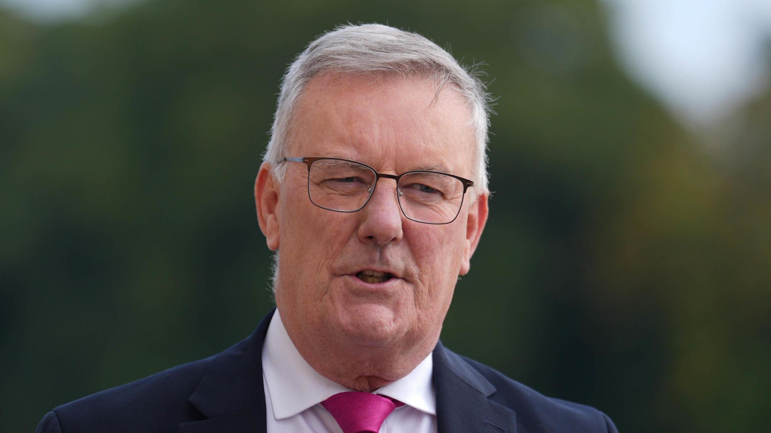 Mike Nesbitt looks to the right of the camera. He has short grey hair and wears clear glasses with black frames, as well ads a navy suit, white shirt and pink tie.