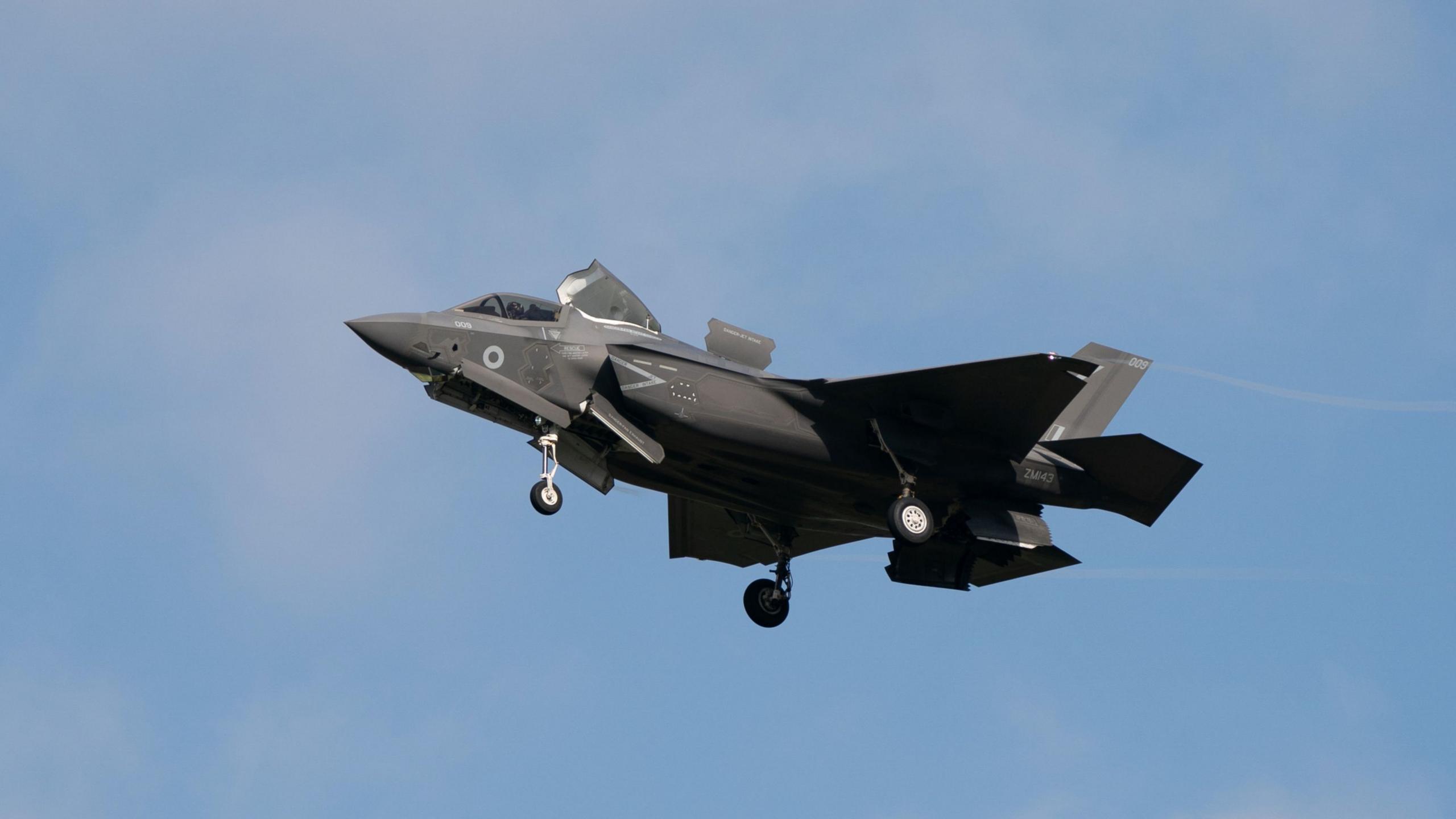 An F35 jet prepares to land against a backdrop of blue sky with faint clouds. The plane is dark grey and has its wheels extended.