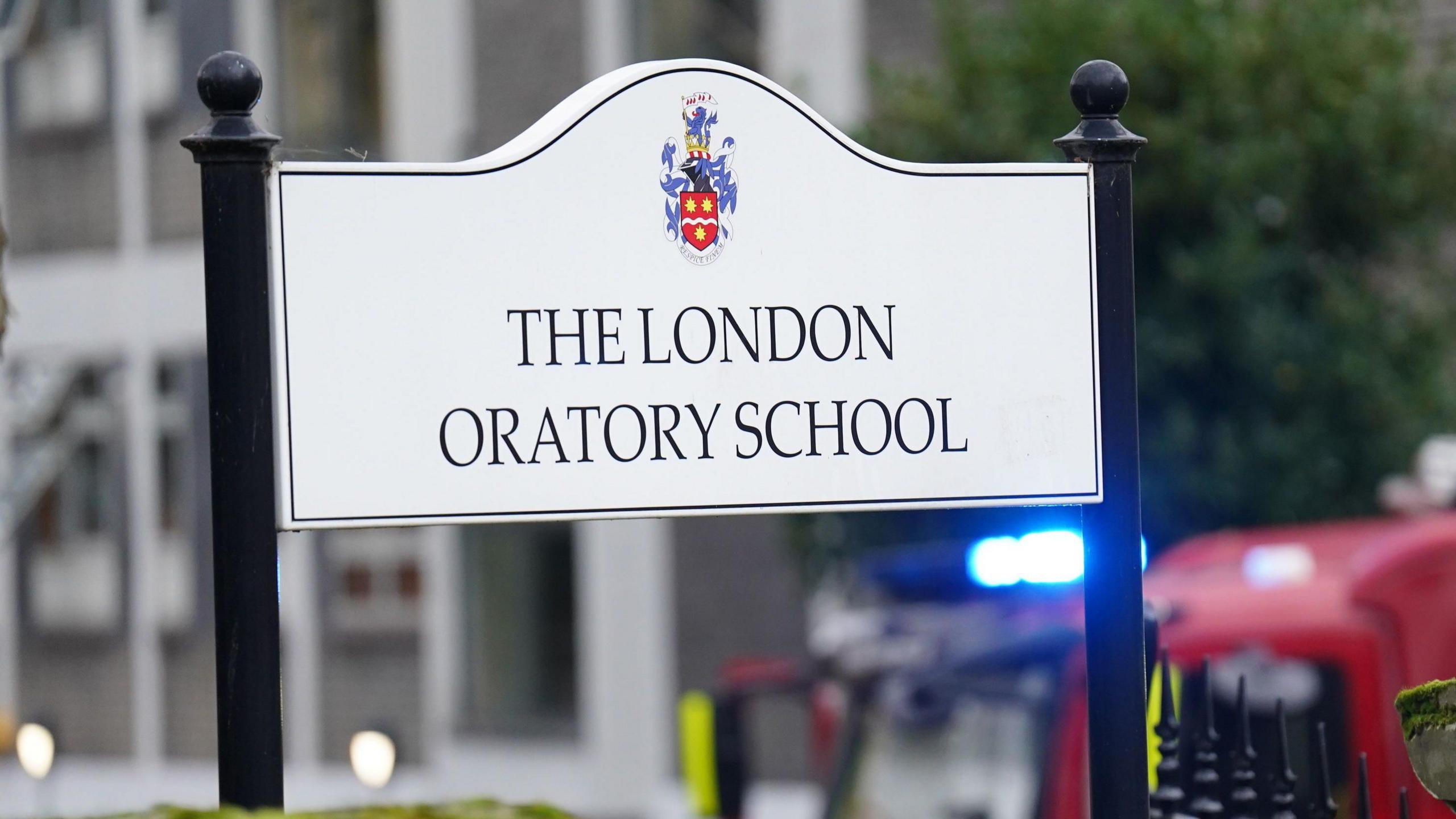 A white sign with "The London Oratory School" in capital letters with the school coat of arms above. In the background is a fire engine with flashing blue lights.  