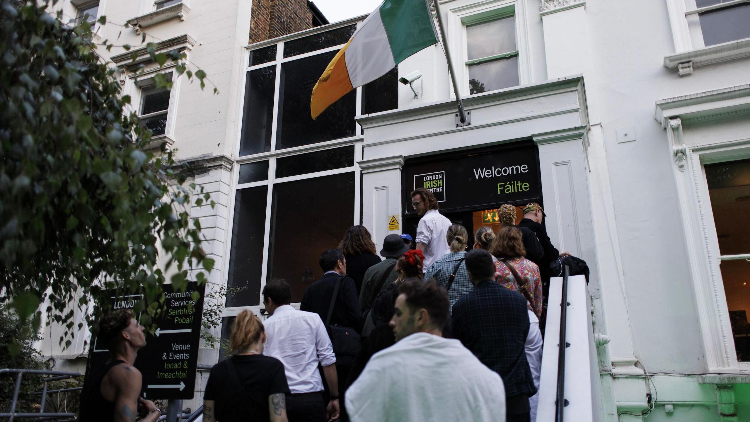 Irish flag flies over the entrance to a white building with black door. More than a dozen people are filing up the steps