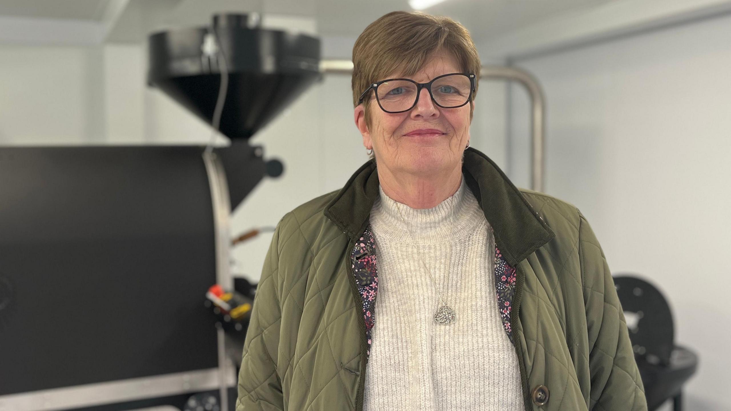 Maureen Flewitt, wearing a green jacket, stands in front of a coffee roasting machine