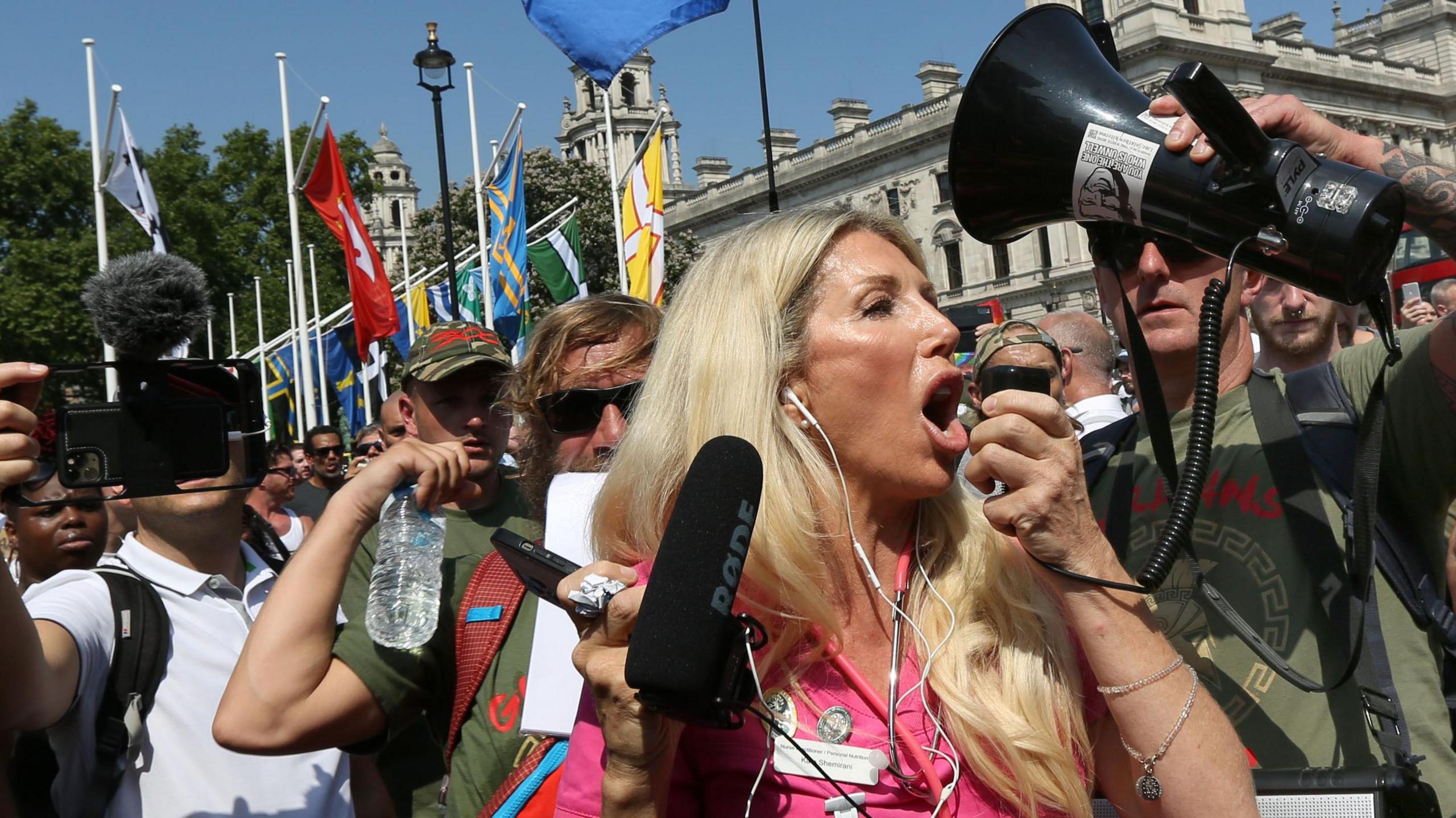 Kate Shemirani, who has long blond hair and wears a pink outfit with what appears to be a stethoscope around her neck, at an anti-lockdown protest in 2021. A man nearby holds up a megaphone speaker as she speaks into the microphone part, which she holds in her hand. Behind her are flags on what looks like Parliament Square, along with crowds of people, some holding up cameras which they are pointing at the press photographer.