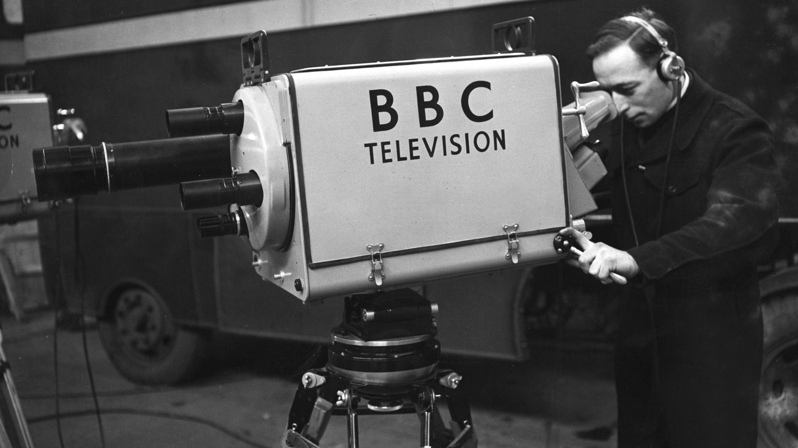A black-and-white shot of a large, old-fashioned television camera, pointing to the left of frame. It has 'BBC Television' written on the side, and has a turret lens set-up, with which to get different focuses a different lens must be physically rotated into place. On the right of frame, a male operator is holding the camera and looking through its viewfinder.
