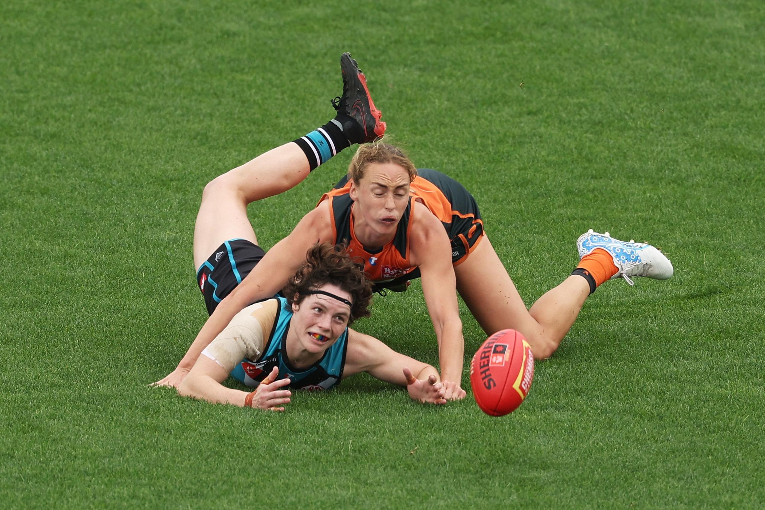 Two players compete for possession of a red football on a grassy field. One player in an orange and black uniform reaches forward while partially pinning the opponent wearing teal and black. The ball lies loose between them during play.