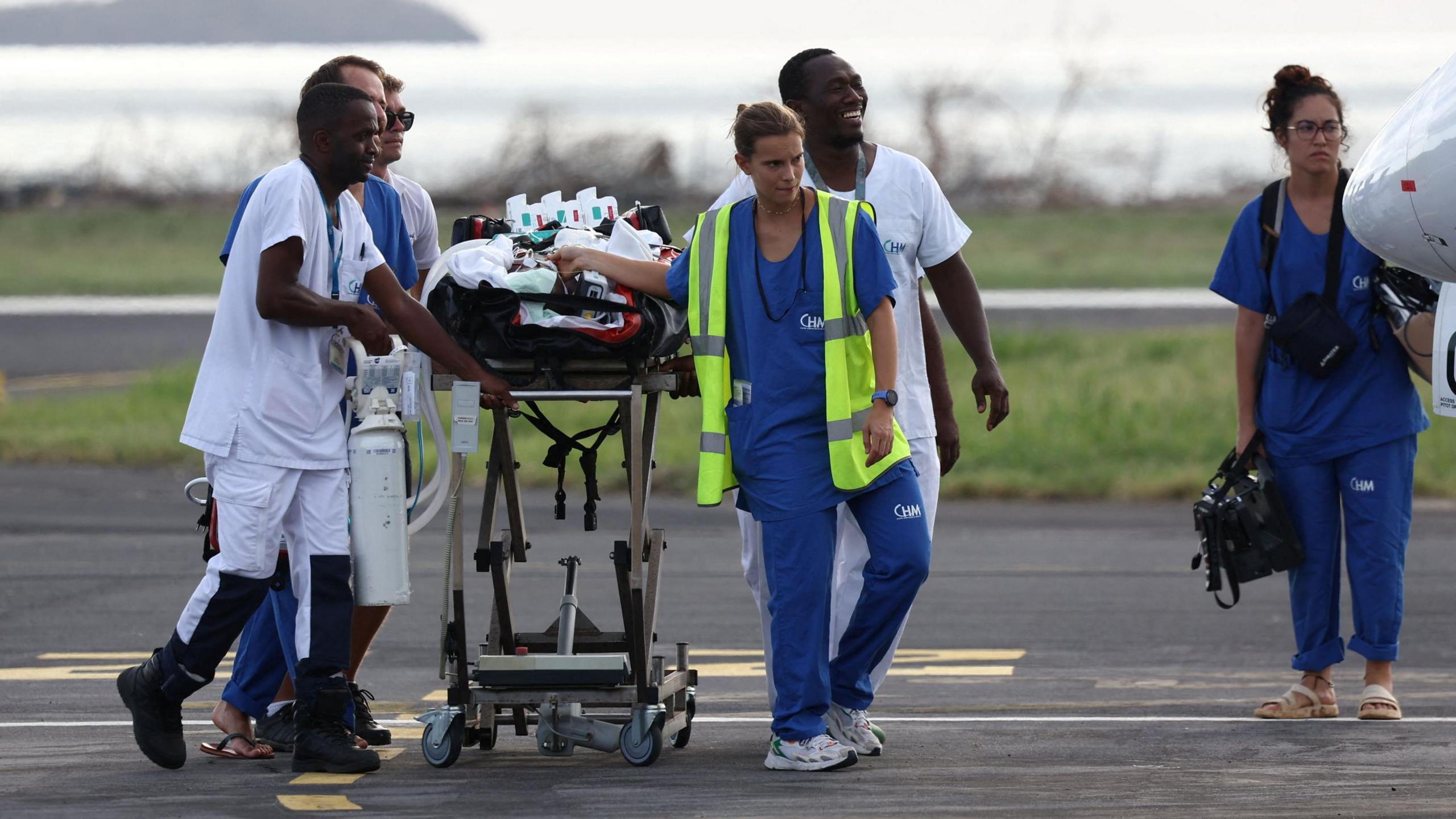 Paramedics in scrubs walk along a runway while pulling a stretcher carrying an injured person to an aircraft