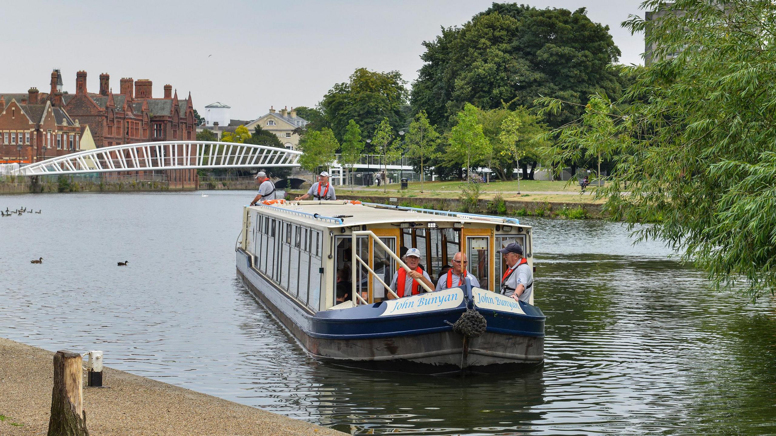 The John Bunyan Community Boat on a trip on the River Great Ouse in Bedford, showing five men on the boat and a bridge and large buildings and greenery in the background.