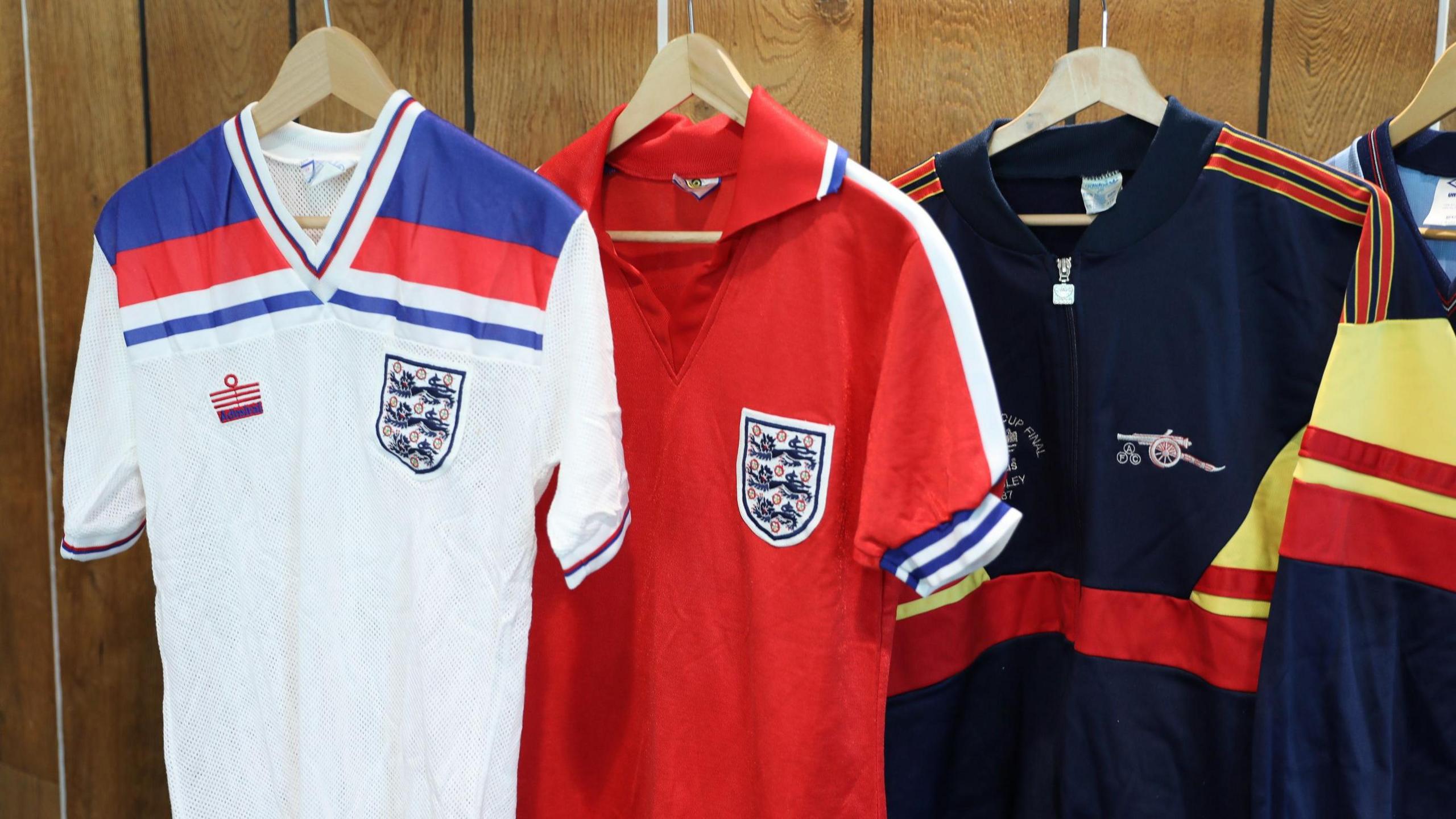 Two of Viv Anderson's England shirts hanging on a clothes rail. The first is white, with an England badge on the left breast, Admiral manufacturer's logo on the opposite side and red and blue horizontal bands across the shoulders. The second is red with an England badge on the left breast and blue and white stripes on the sleeve. Alongside them is a dark blue Arsenal tracksuit from the 1987 Littlewoods Cup Final. It has yellow and red detailing.