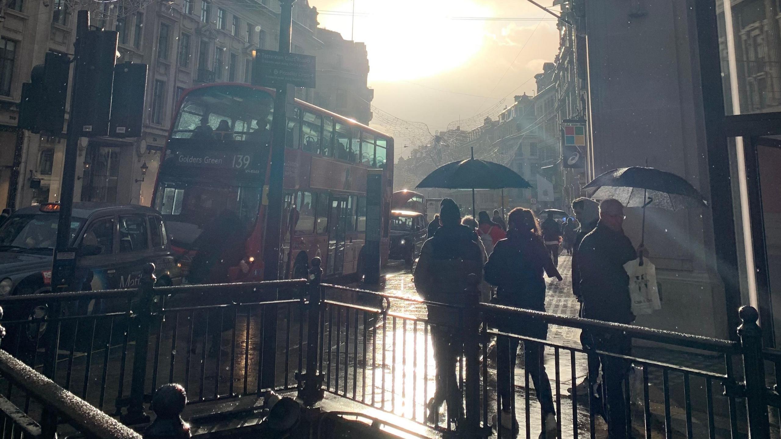 A busy London street scene at Oxford Circus, where sunshine breaks through passing showers, illuminating wet pavements, with pedestrians carrying umbrellas. 