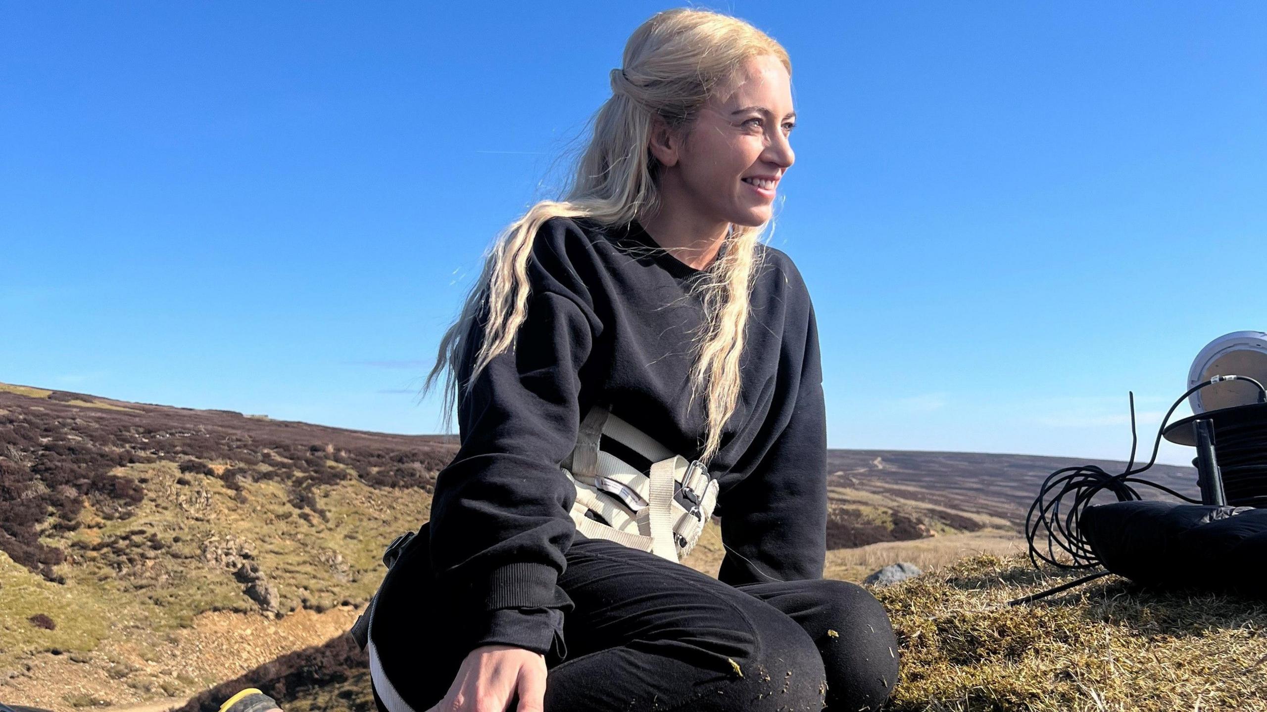 Nikita Mitchell mban një parzmore mbi një bluzë blu. Ajo ka flokë të gjatë bjondë dhe është ulur në një kodër duke parë në distancë.