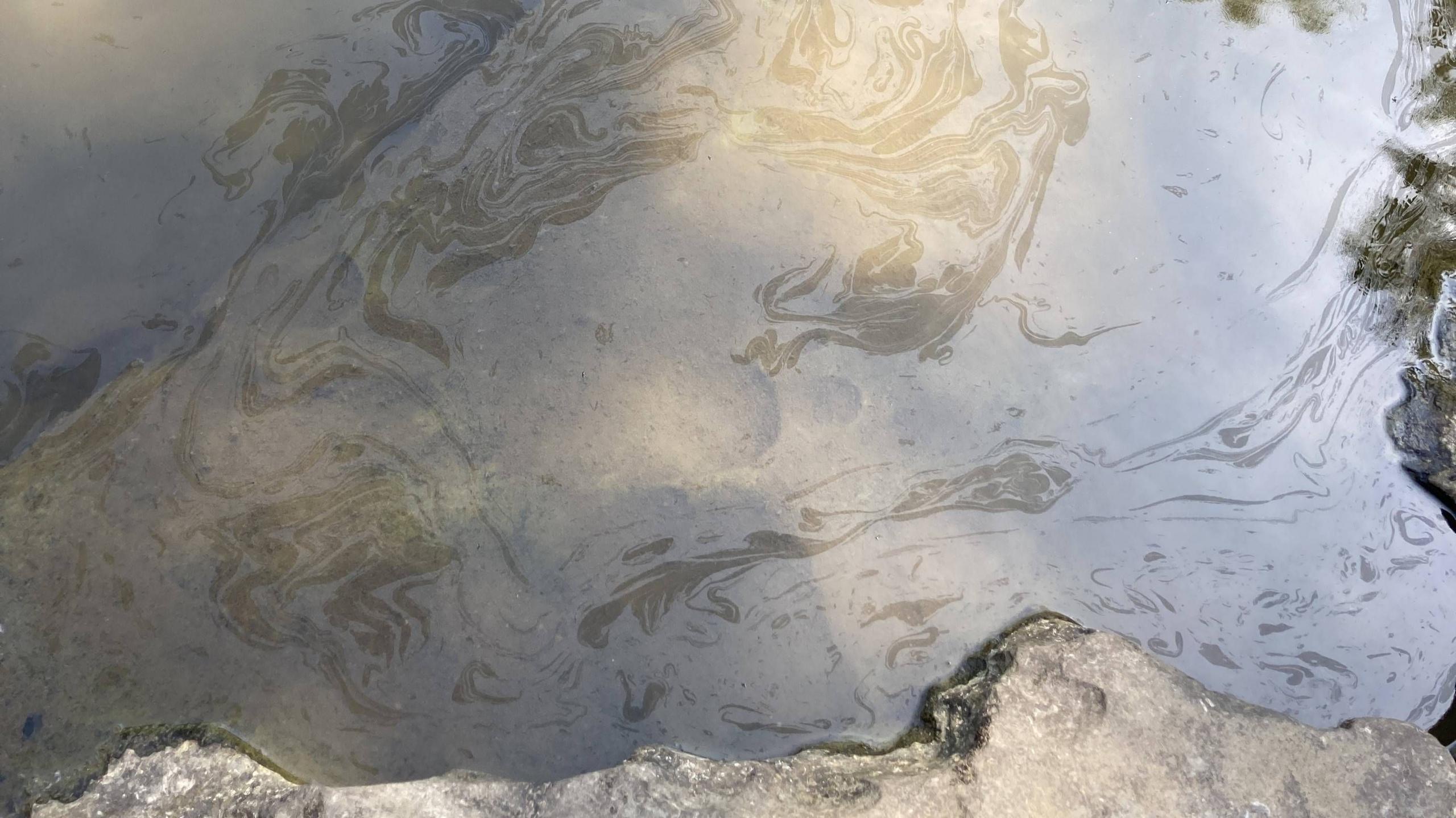 The surface of a watercourse is oily, with swirls through the water. Rocks at the side of the beck are visible at the bottom of the frame.