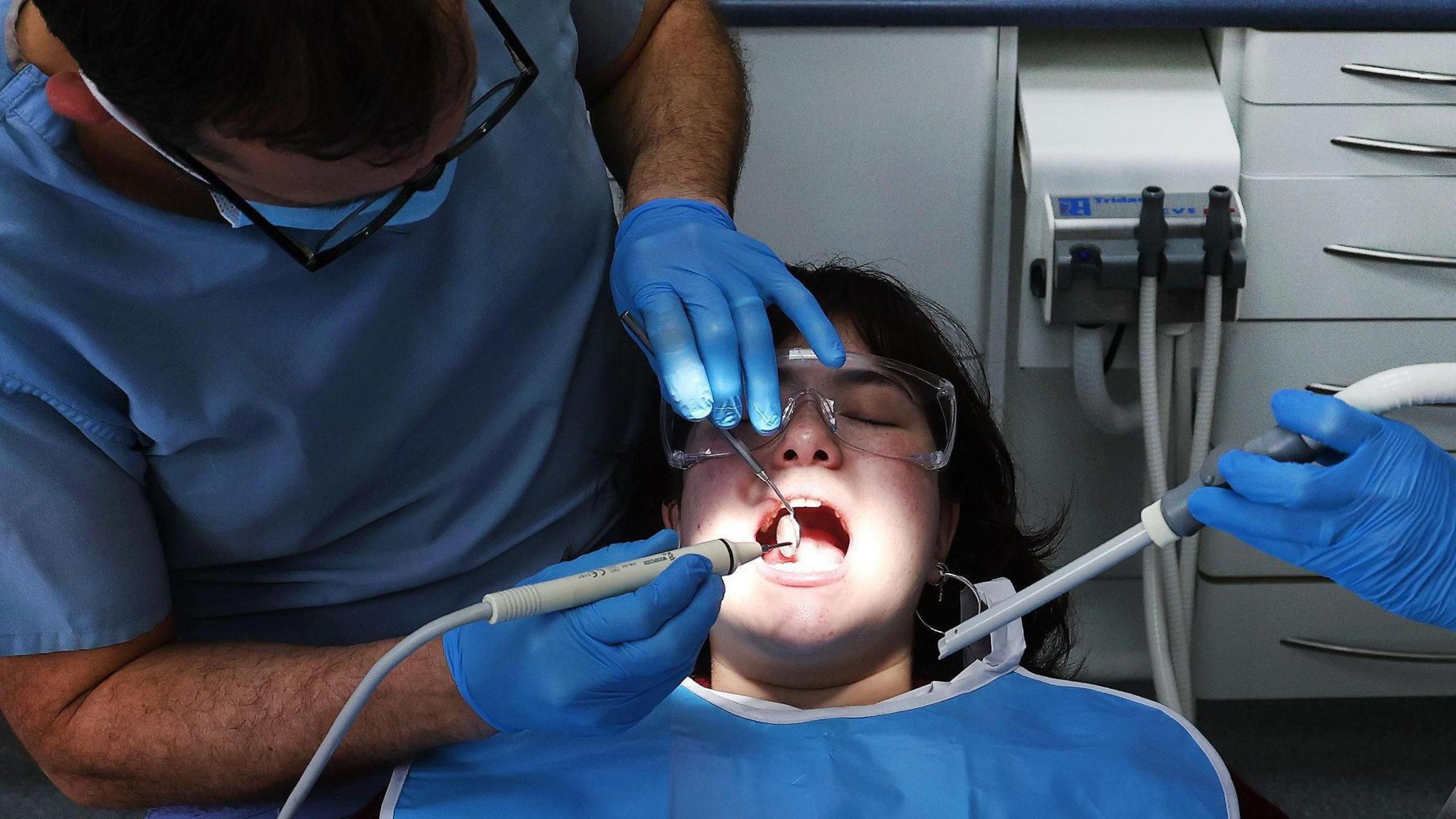 A patient lies back on a dentist's chair with her mouth open and wearing a blue apron. A dentist standing to her left is examining her mouth with tools whilst wearing a pair of blue gloves and glasses. To the patient's right is a dental nurse in a red set of scrubs, also wearing blue gloves, she is holding a suction hose below the patient's face.