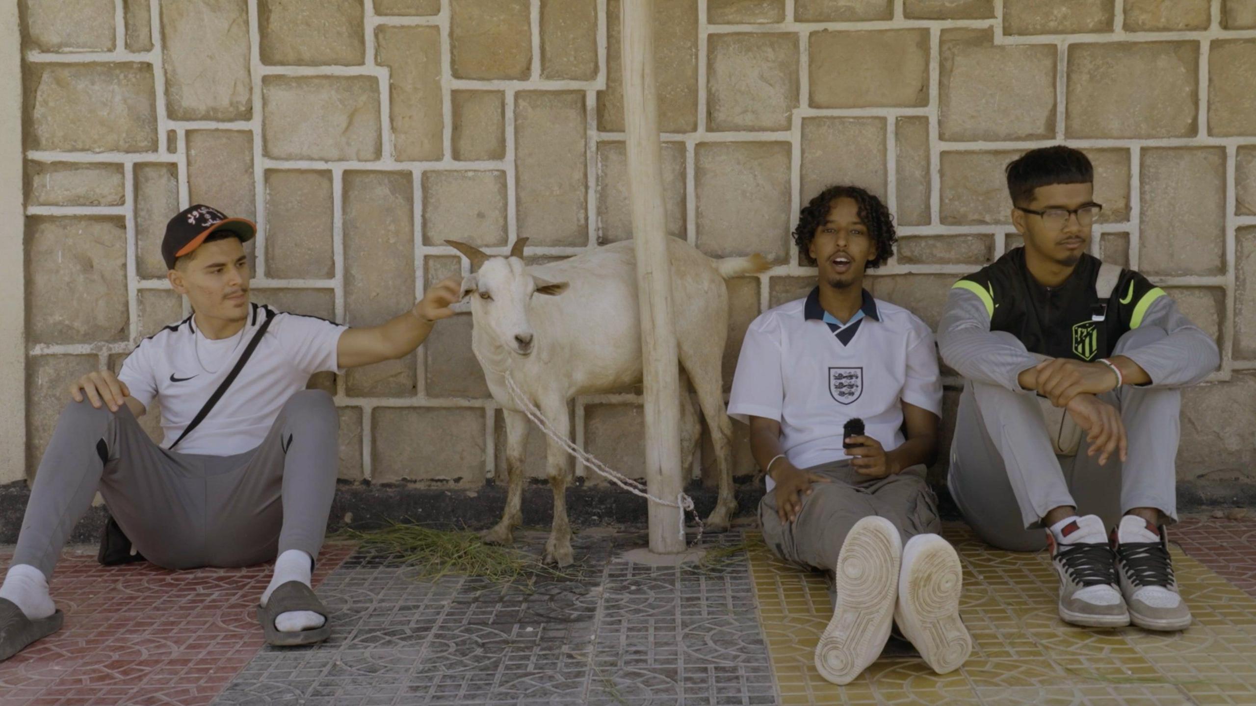 Zak Hajjaj, Abu Finiin, Kayum Miah sitting down against a wall with Zak petting a goat between himself and Abu