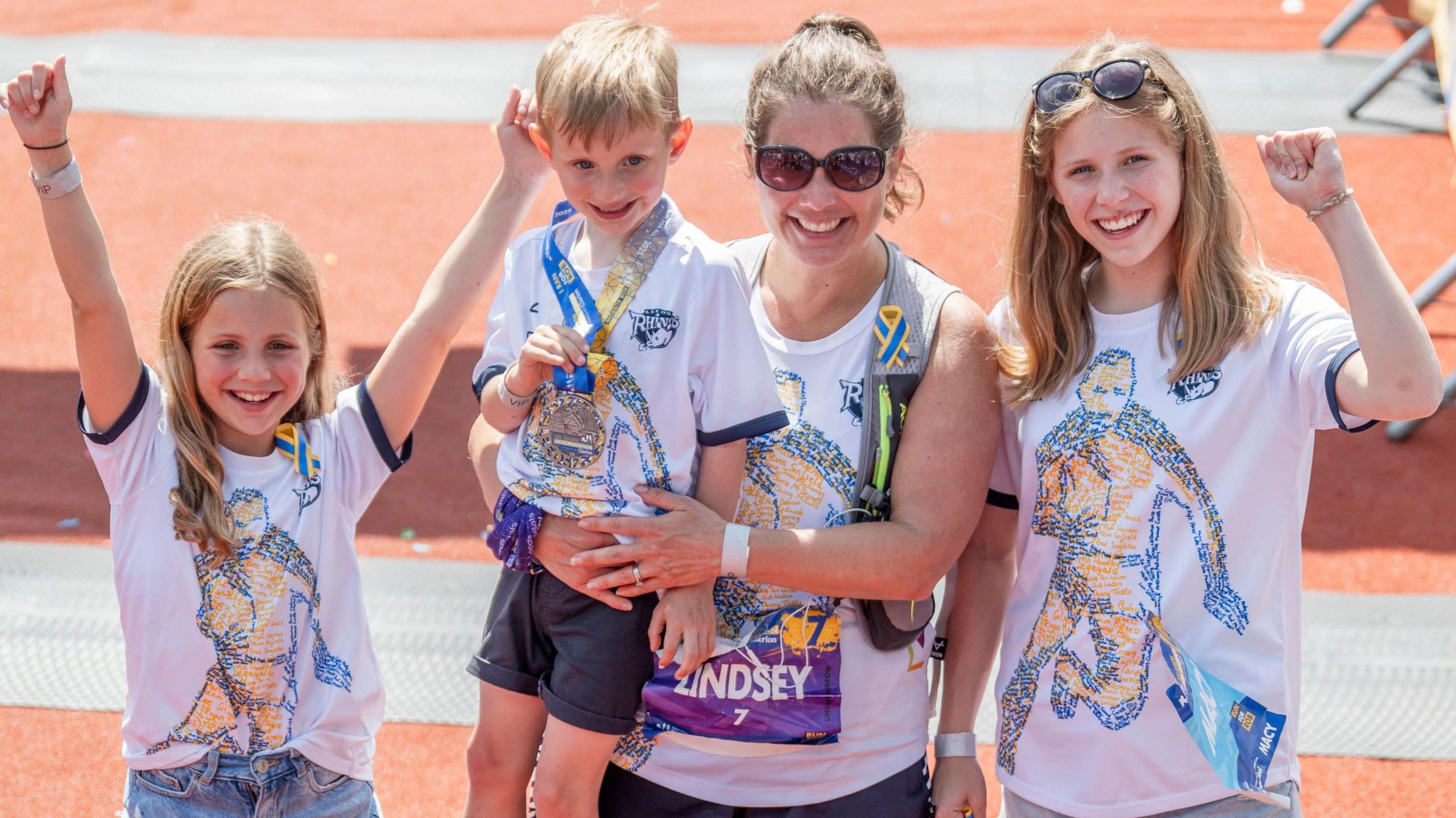 Lindsey wears running shorts and sunglasses and has her hair in a ponytail. She is holding her son in her arm. Either side of her are girls who look about 10 and 13, all cheering. They all have matching white T-shirts with Rob on.