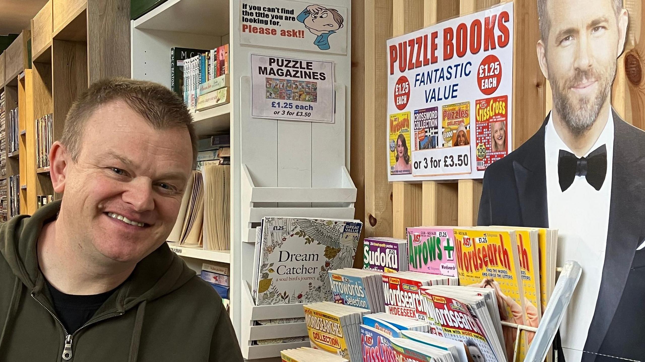 Rob Clarke smiling at the camera stood in his DVD shop next to a cardboard cut-out of Ryan Reynolds in a suit. A magazine rack is full of puzzle magazines to his life.