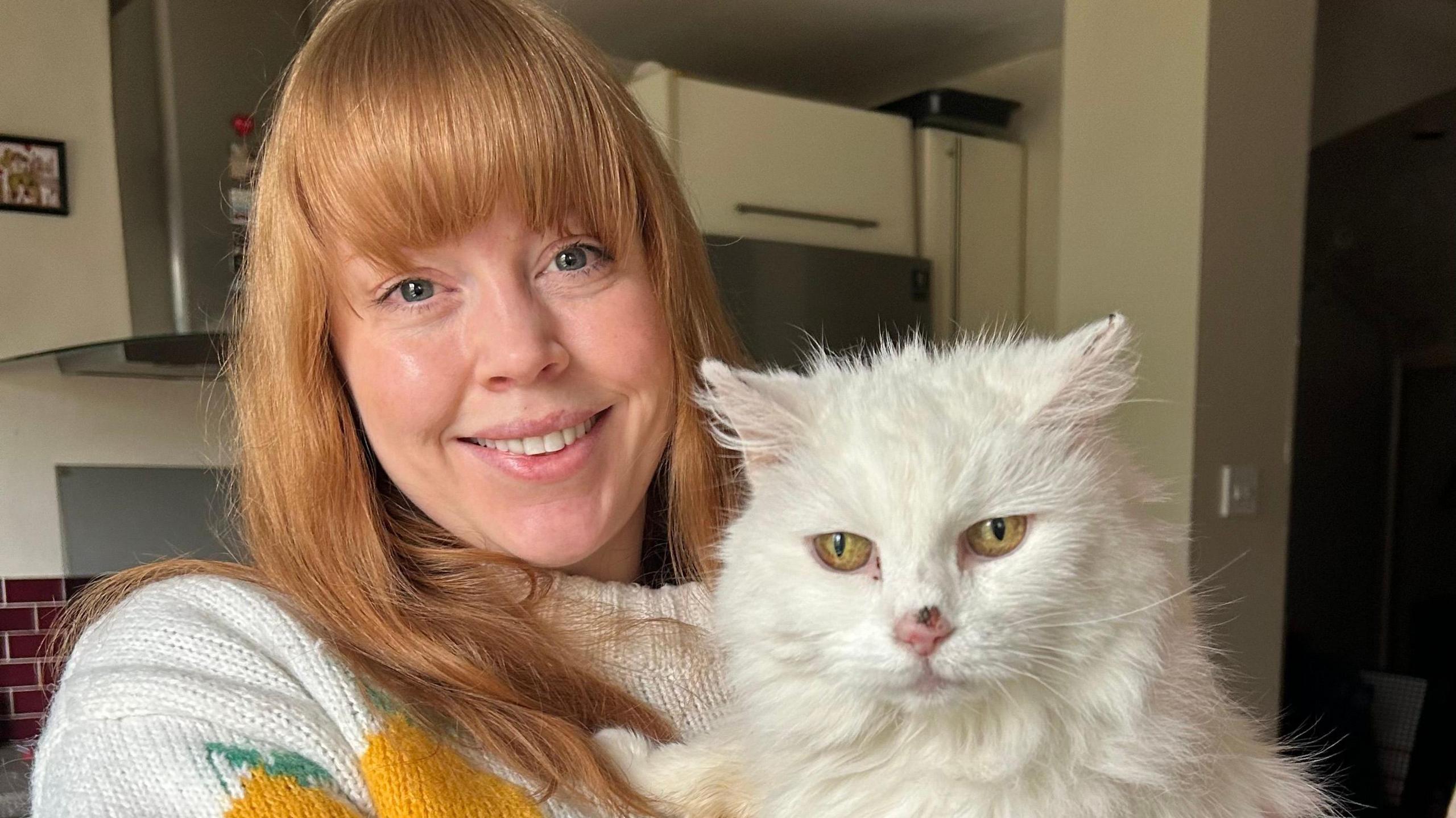 Bex Hunt holding Pixie, a white cat, they are both looking at the camera, Bex is smiling, with long hair and fringe, a white jumper, there are kitchen cabinets behind her. 