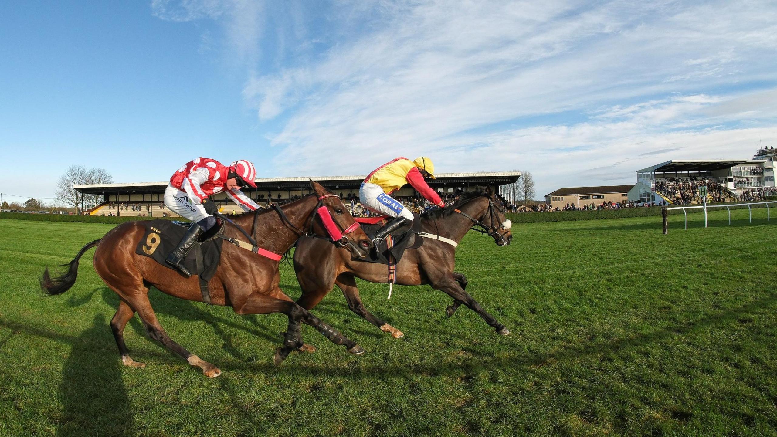 Two jockeys race down the home straight on their horses at Wincanton Racecourse in Somerset. One of them is Harry Kimber riding My Mate Polly. He is wearing a red top with large white spots. Two grandstands containing spectators are visible in the background