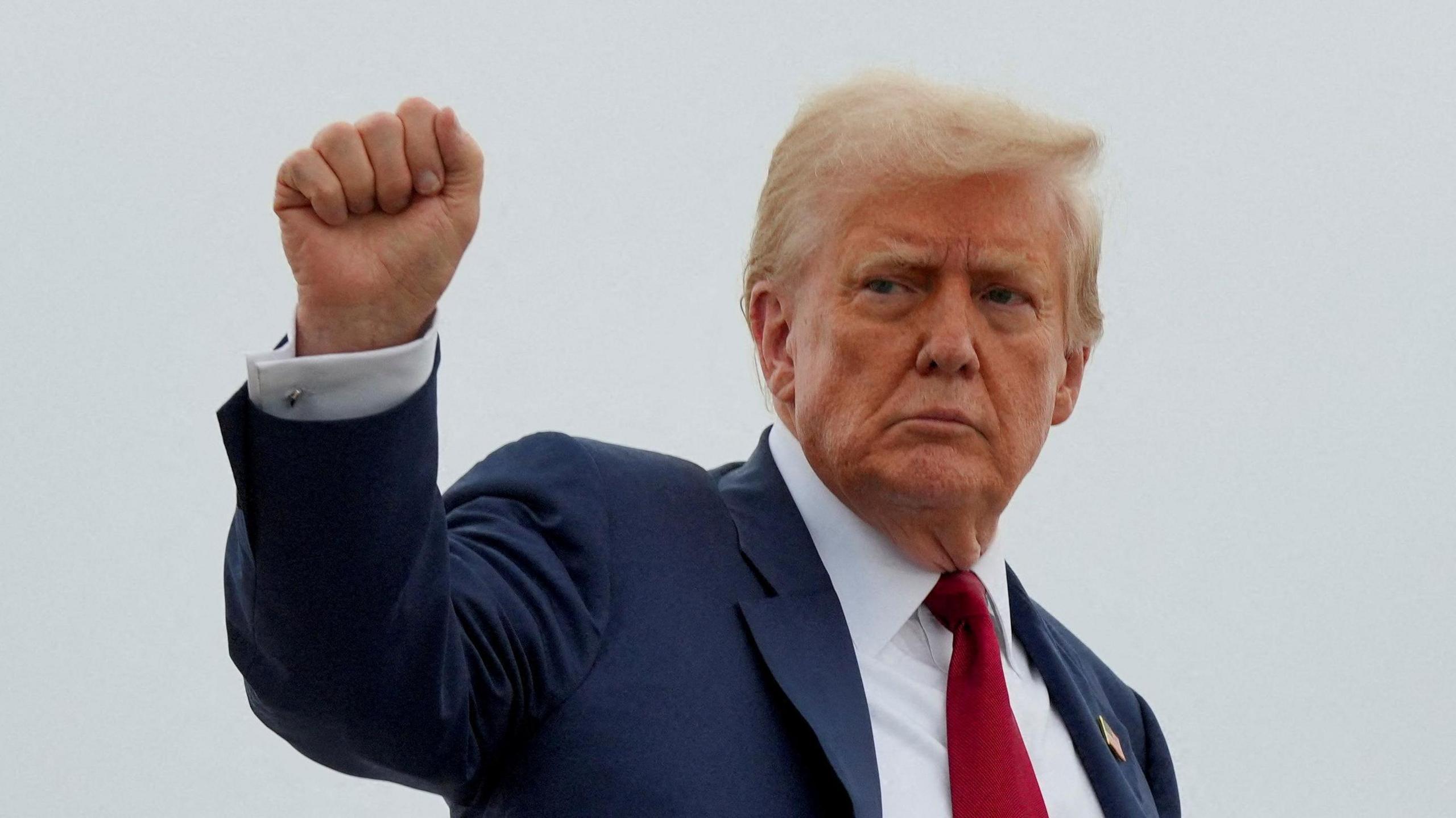 Donald Trump wears a blue suit, white shirt and red tie. He stands in front of a cloudy grey sky and raises his right hand in a fist.