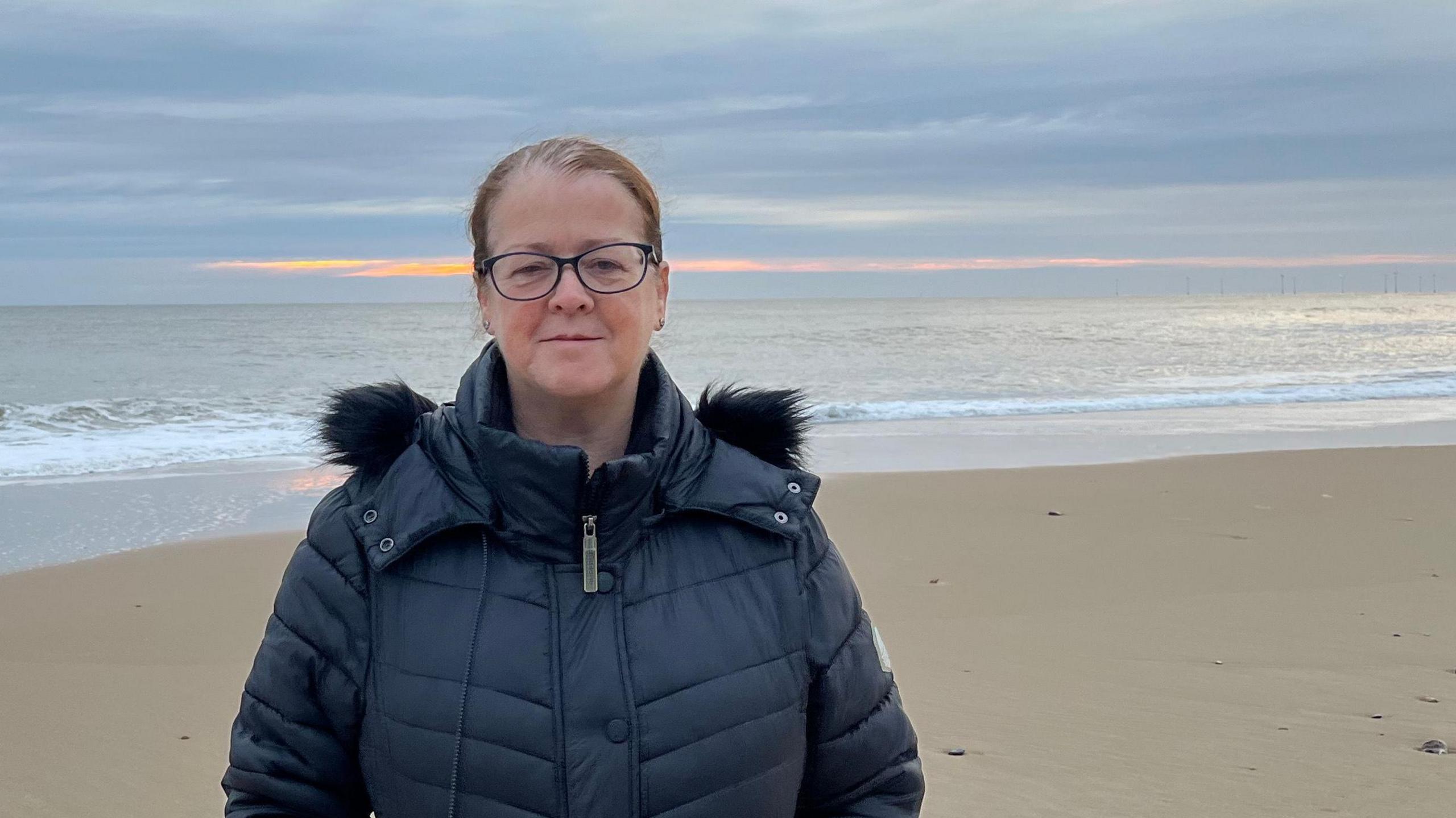Jenny Measures who has pulled back dark hair and is wearing black-framed glasses and a purple jacket. Behind her is a sandy beach, a calm sea and a sky with a streak of yellow and pink from the rising sun. 