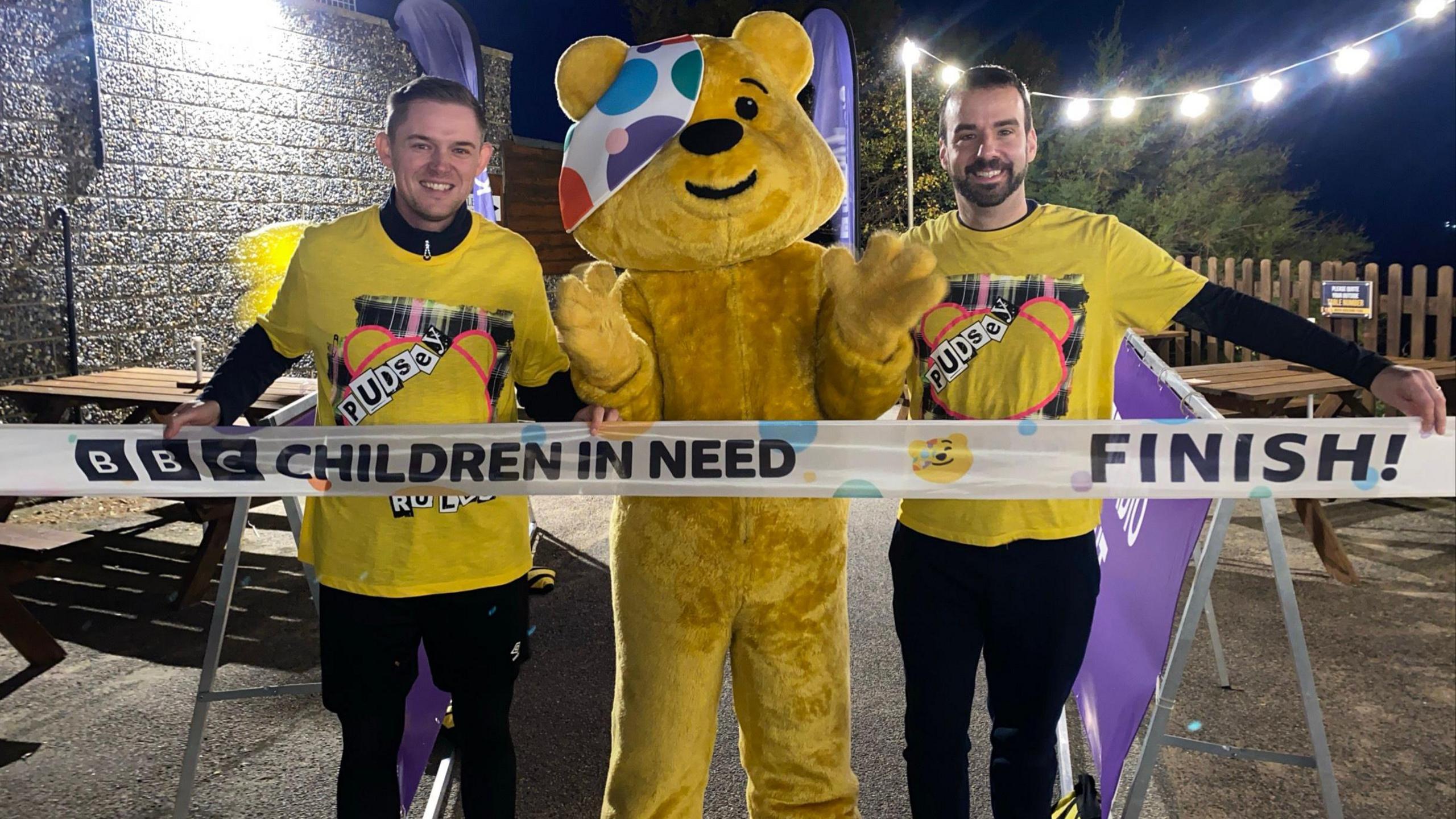 Two men in yellow shirts and black trousers standing beside a big yellow mascot bear. In front of them is a finishing line tape.