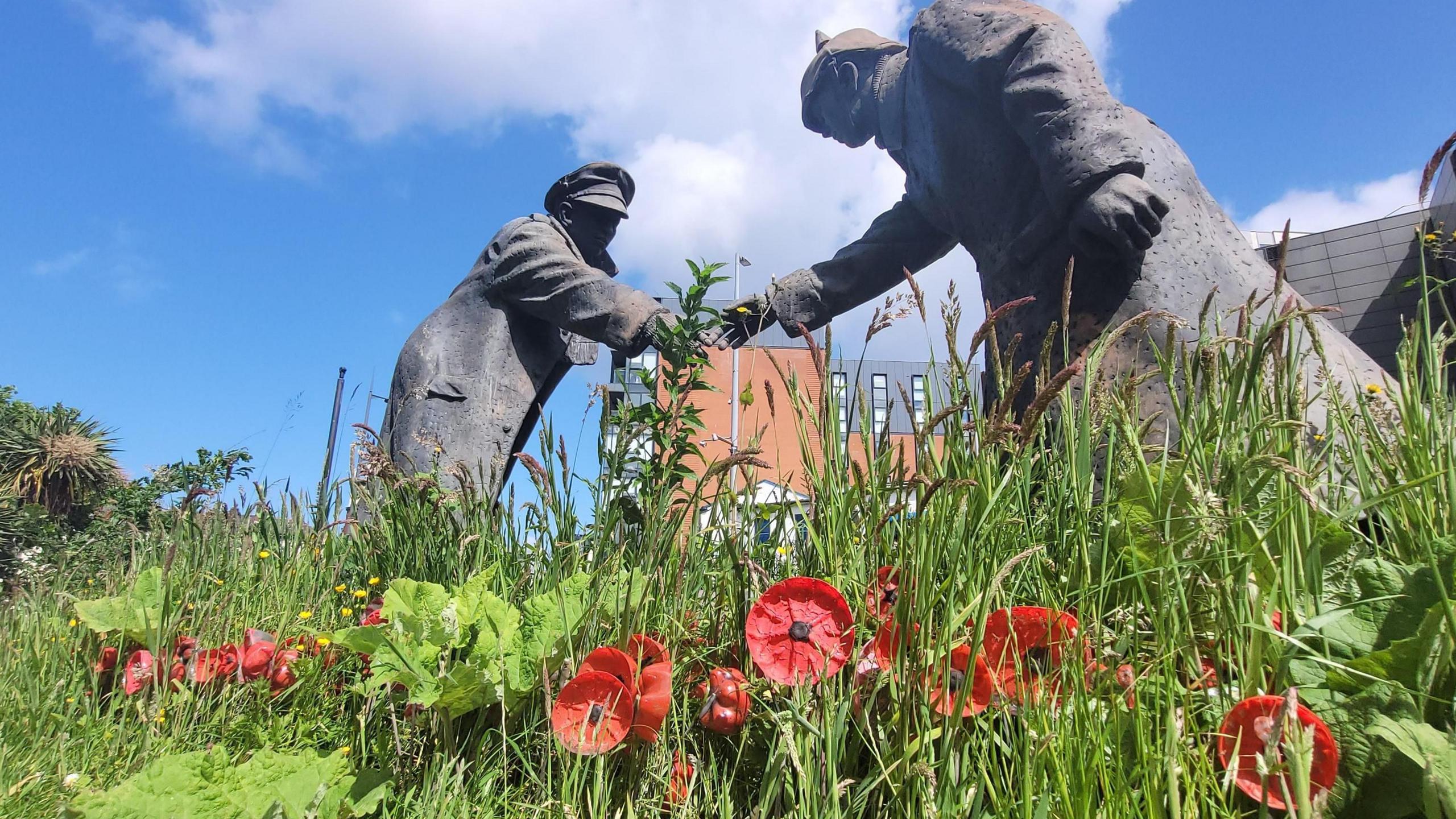 War memorial with two soldiers reaching to shake hands. Below them is a field of commemorative poppies, with some blue sky and light cloud overhead