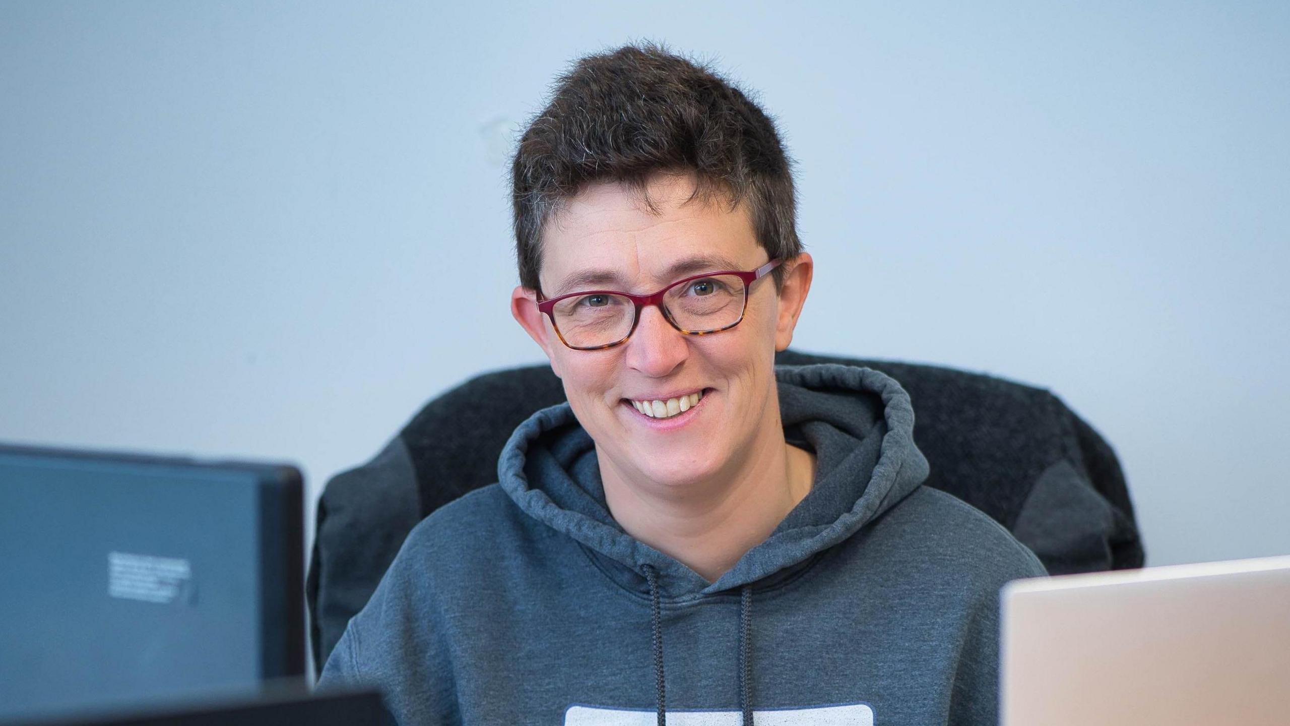 A woman with short dark hair and wearing a grey hoodie, sitting in a chair in an office, with a laptop computer sitting in front of her.