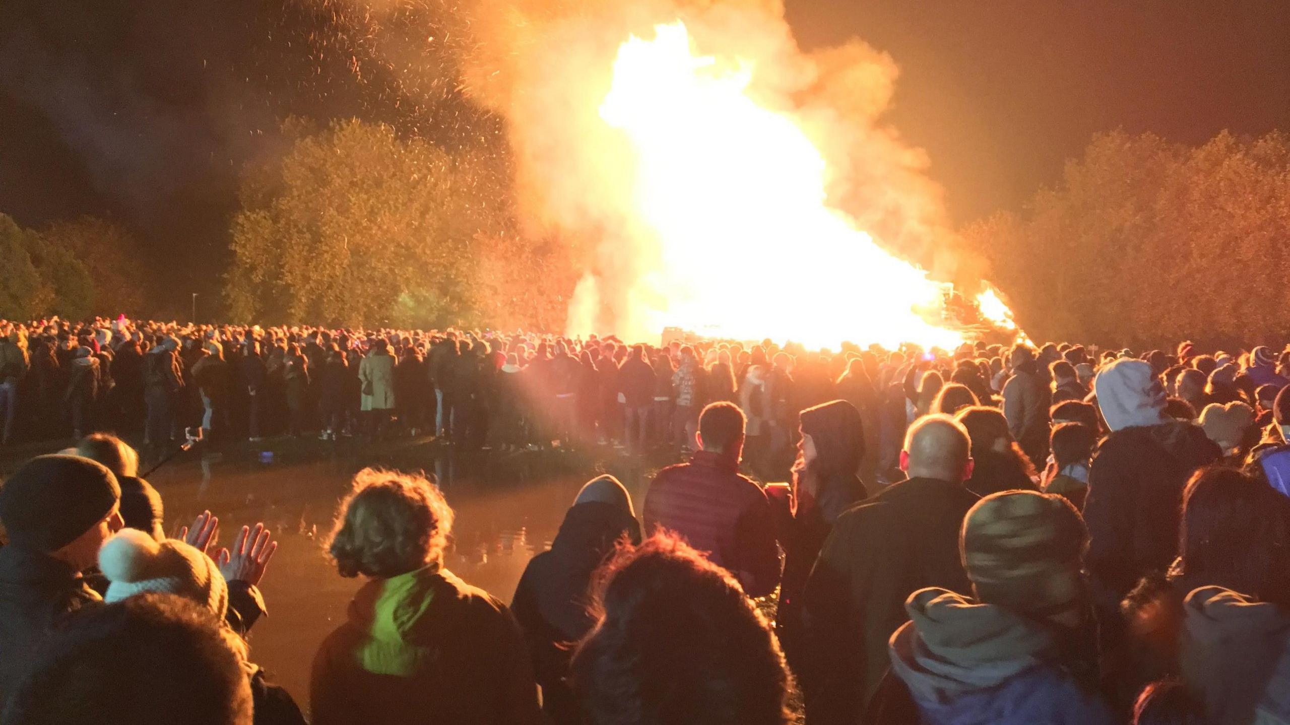 A large group of people standing around the Lewes Bonfire watching the flames leap into the sky.