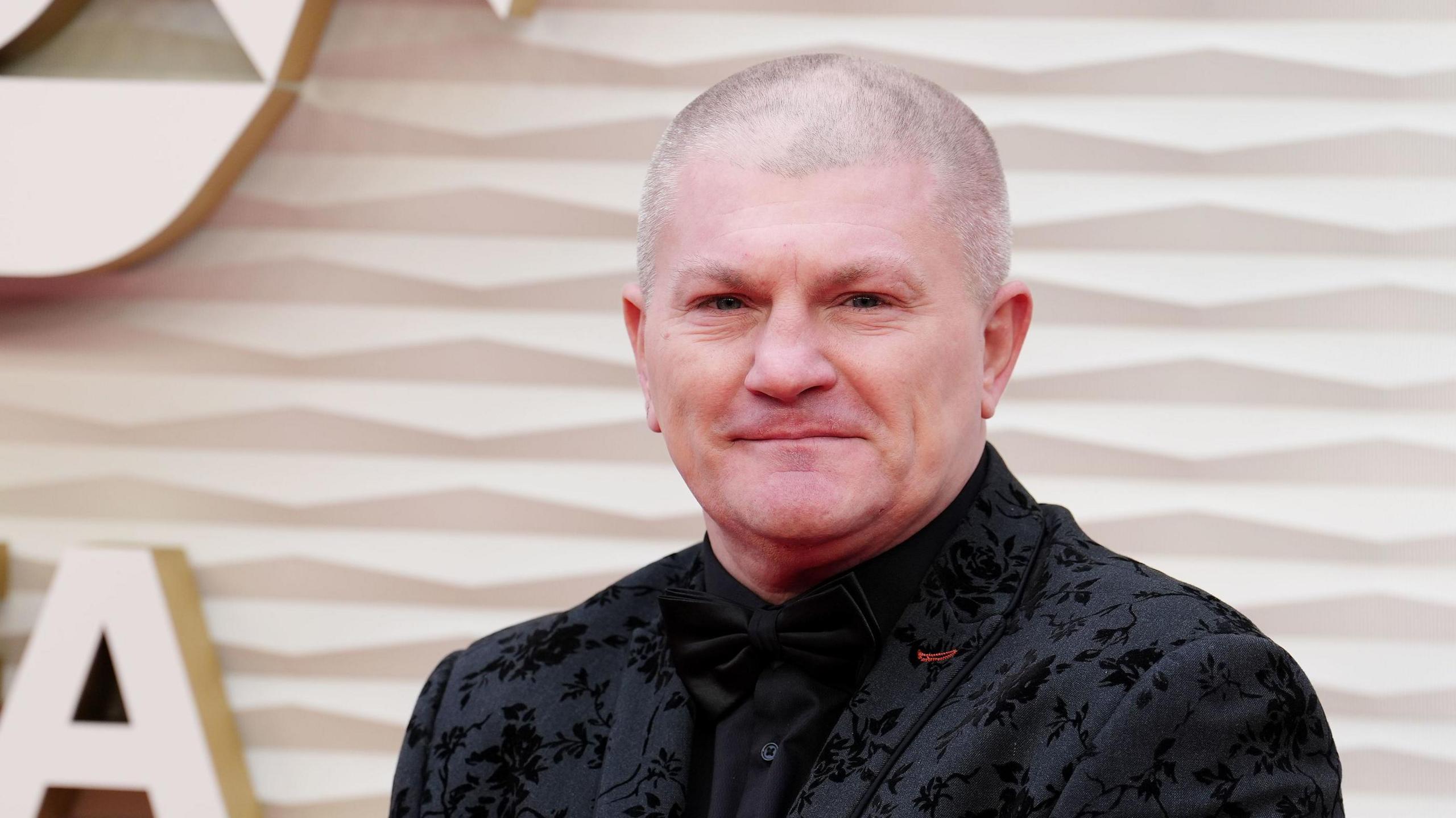 Ricky Hatton arrives for the PFA Awards 2025. He has a shaven head and wears a black embroidered suit with black shirt and black bow tie as he smiles for the cameras. He is against a golden backdrop.