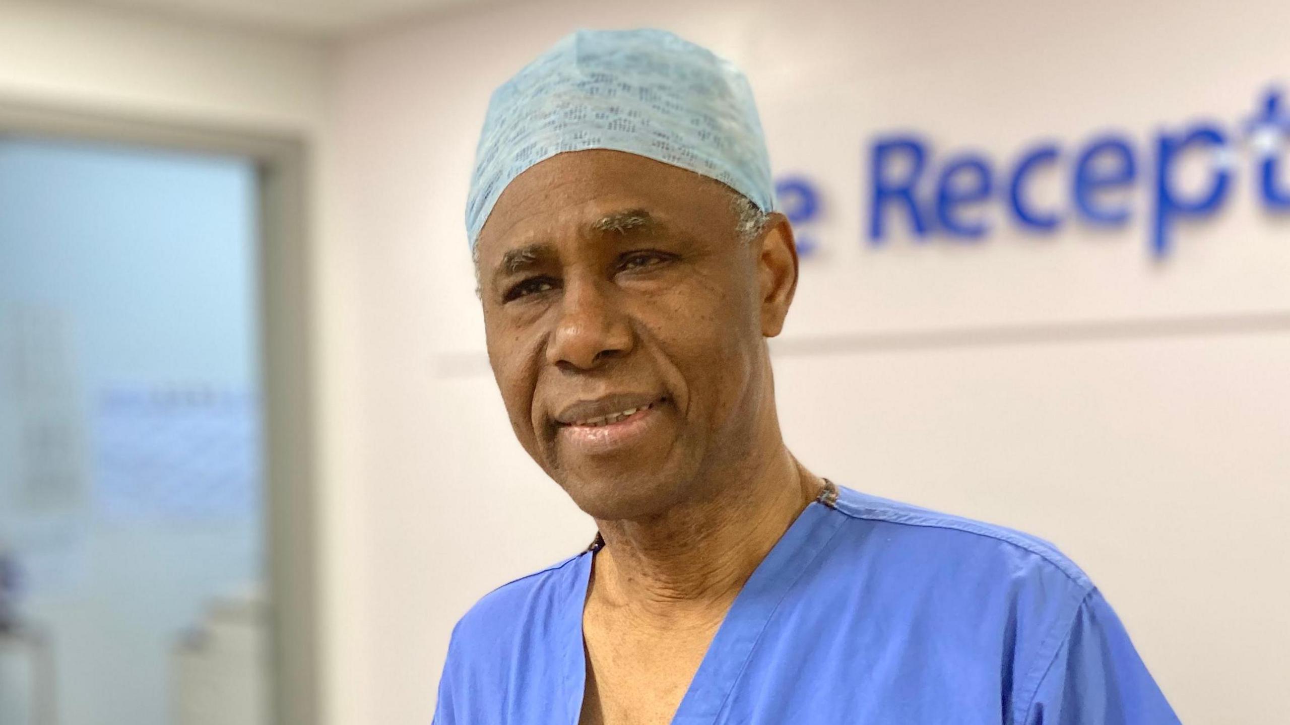 Mr Akomolafe is stood in front of a hospital reception desk. He is wearing theatre scrubs, which includes a light blue surgical cap and blue top.