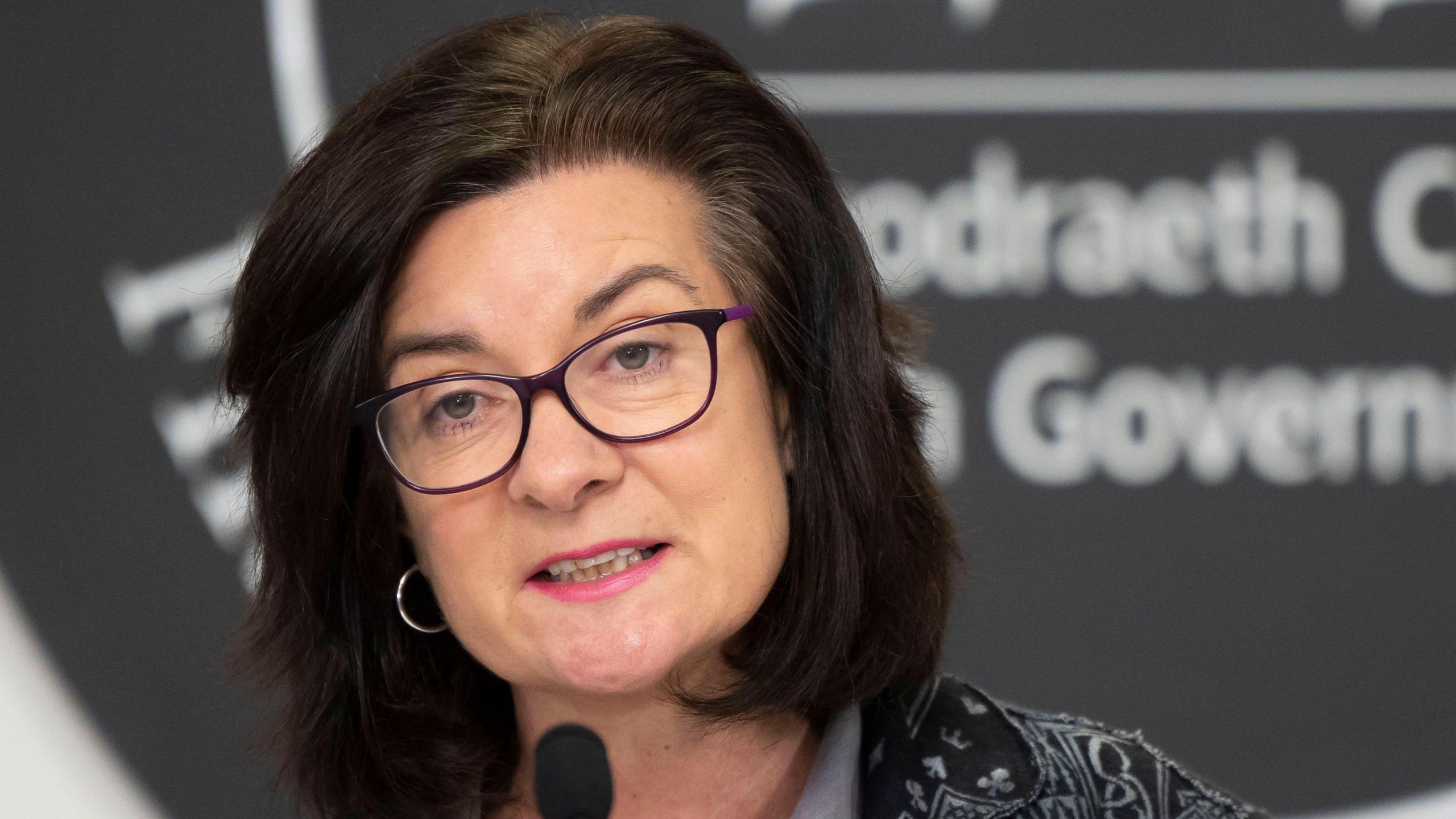 Eluned Morgan speaking at a Welsh government press conference, she is looking at the camera and wearing purple glasses and pink lipstick