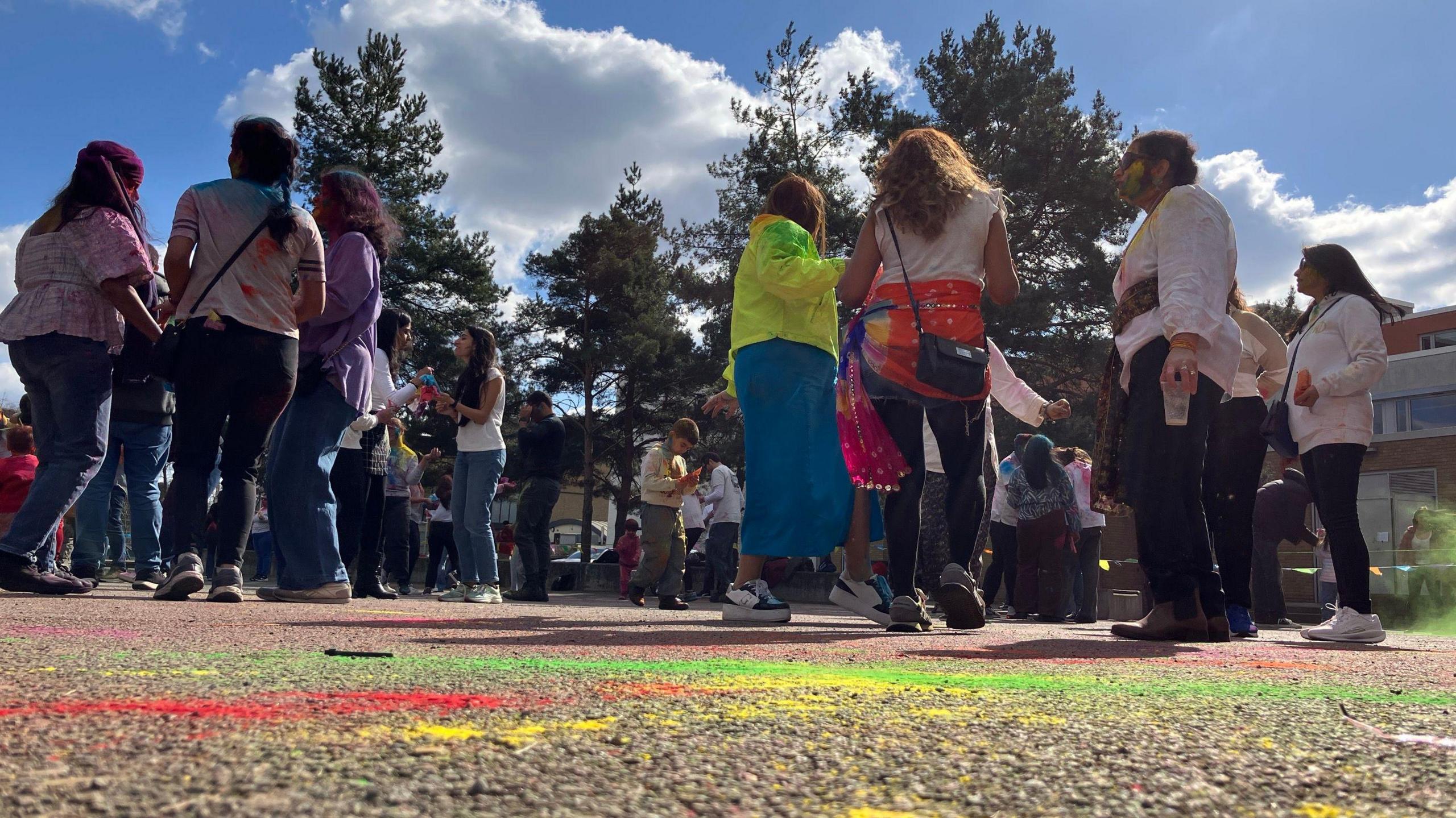 Groups of people stand around chatting - on the ground are colourful streaks of green, red and yellow powder.