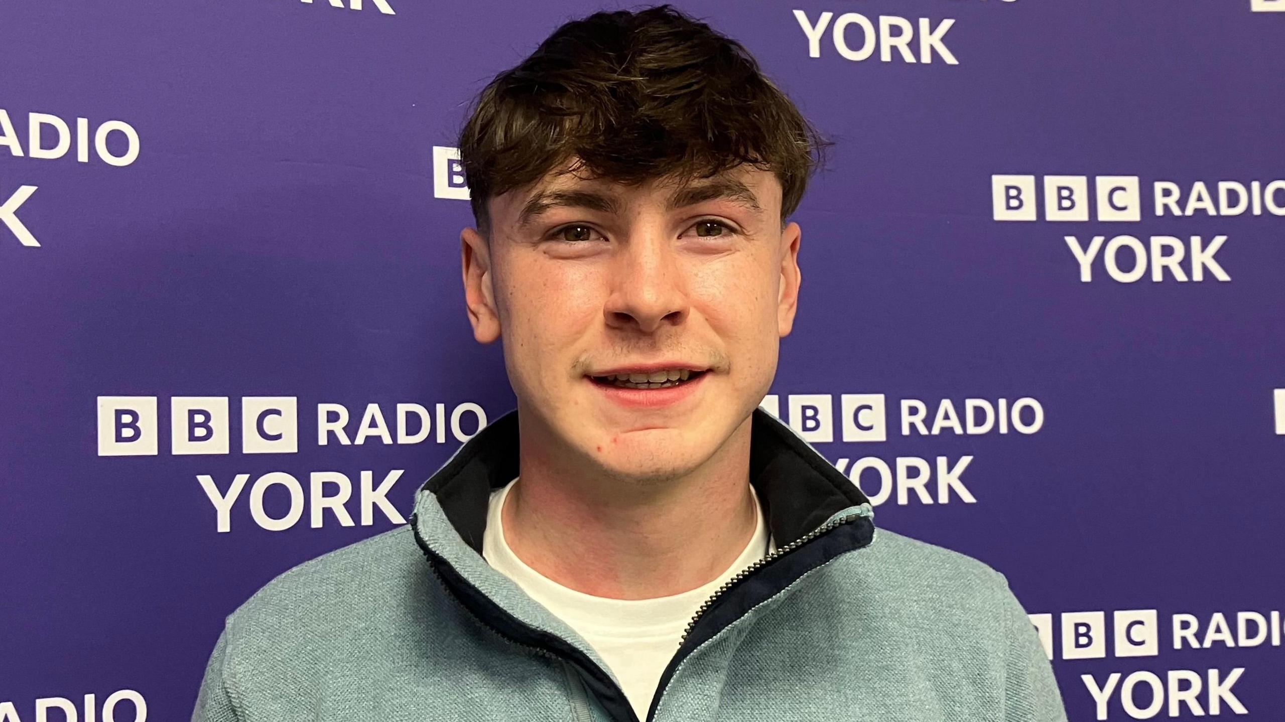 A young man stands in front of a purple BBC Radio York backdrop featuring repeated white text. He wears a light blue quarter-zip pullover over a white shirt.