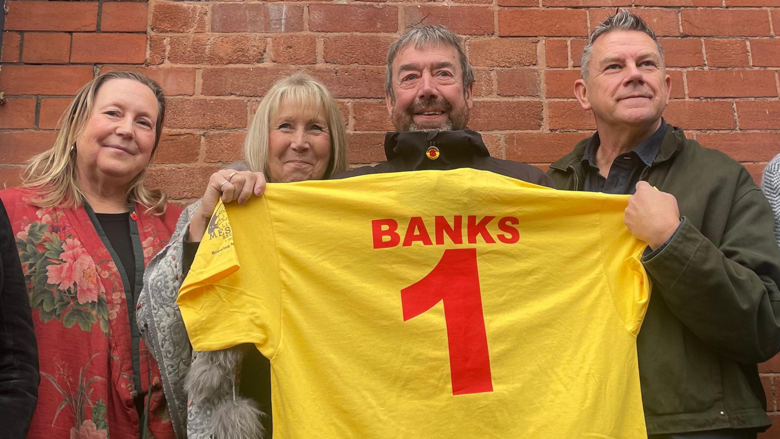 Two men and two women stand in a line holding up a yellow football shirt with the number one in red printed on it