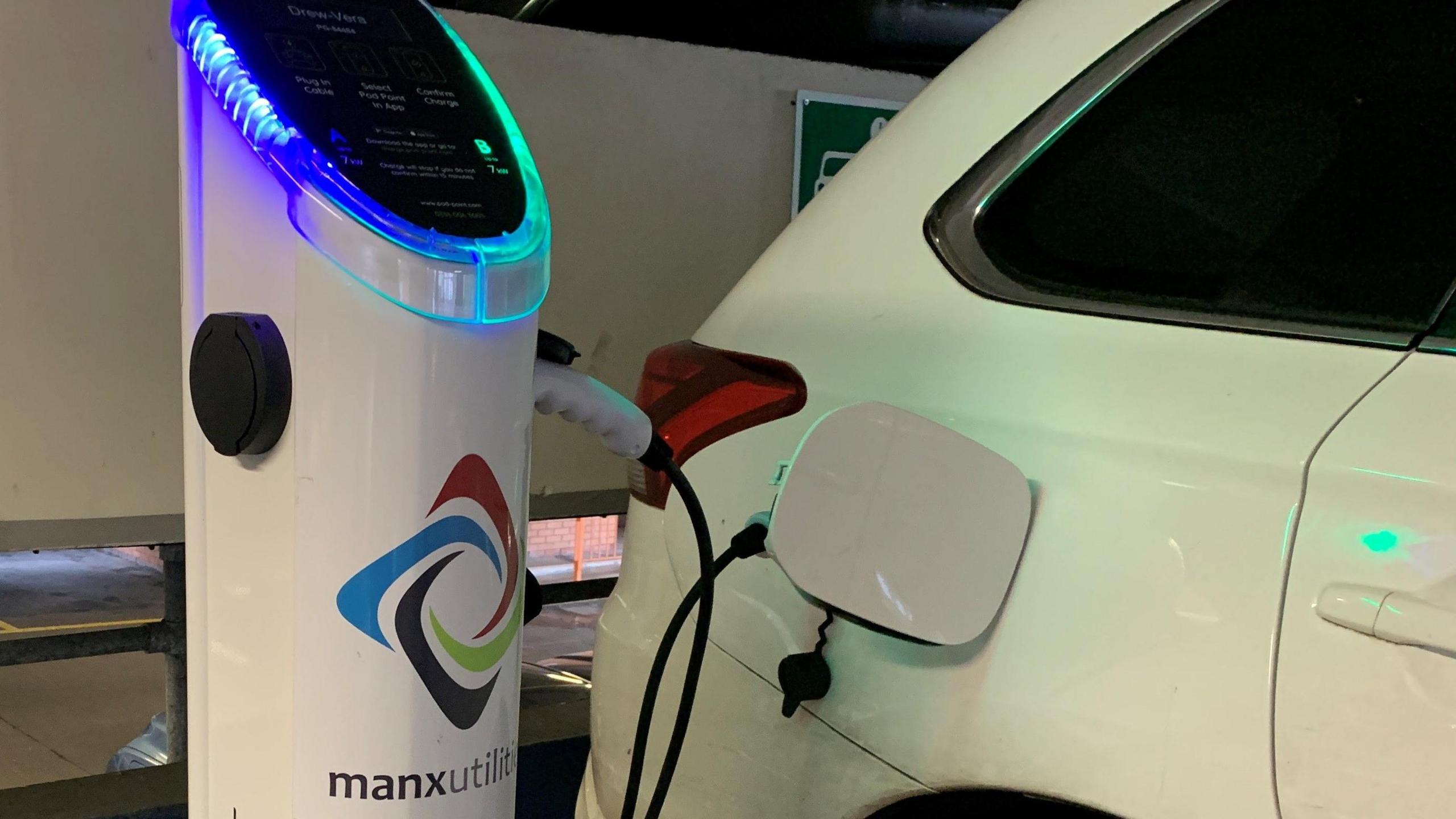 A white car parked next to a charging point in a multi-storey car park. The charging point in a the charging point is white the the Manx Utilities logo on the front and has a black screen on the top. A cable is plugged in connecting the charging point and the car.