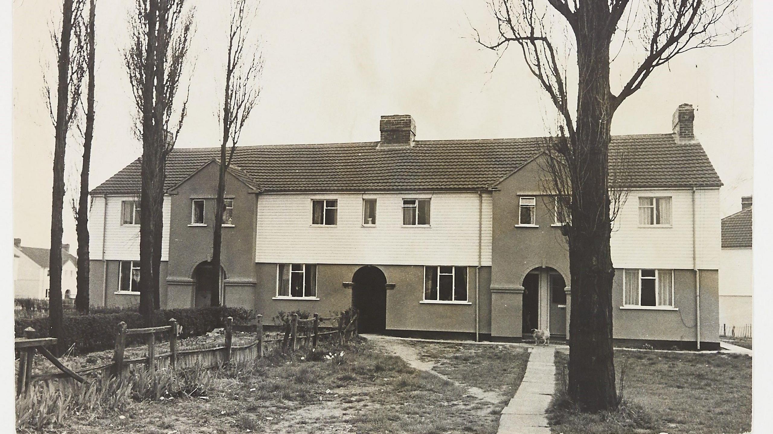 A black and white photo of terraced homes on two storeys. The homes are white at the top with brickwork on the bottom storey. They also have chimneys. In front of the homes are several trees and some grass areas with a wooden fence.