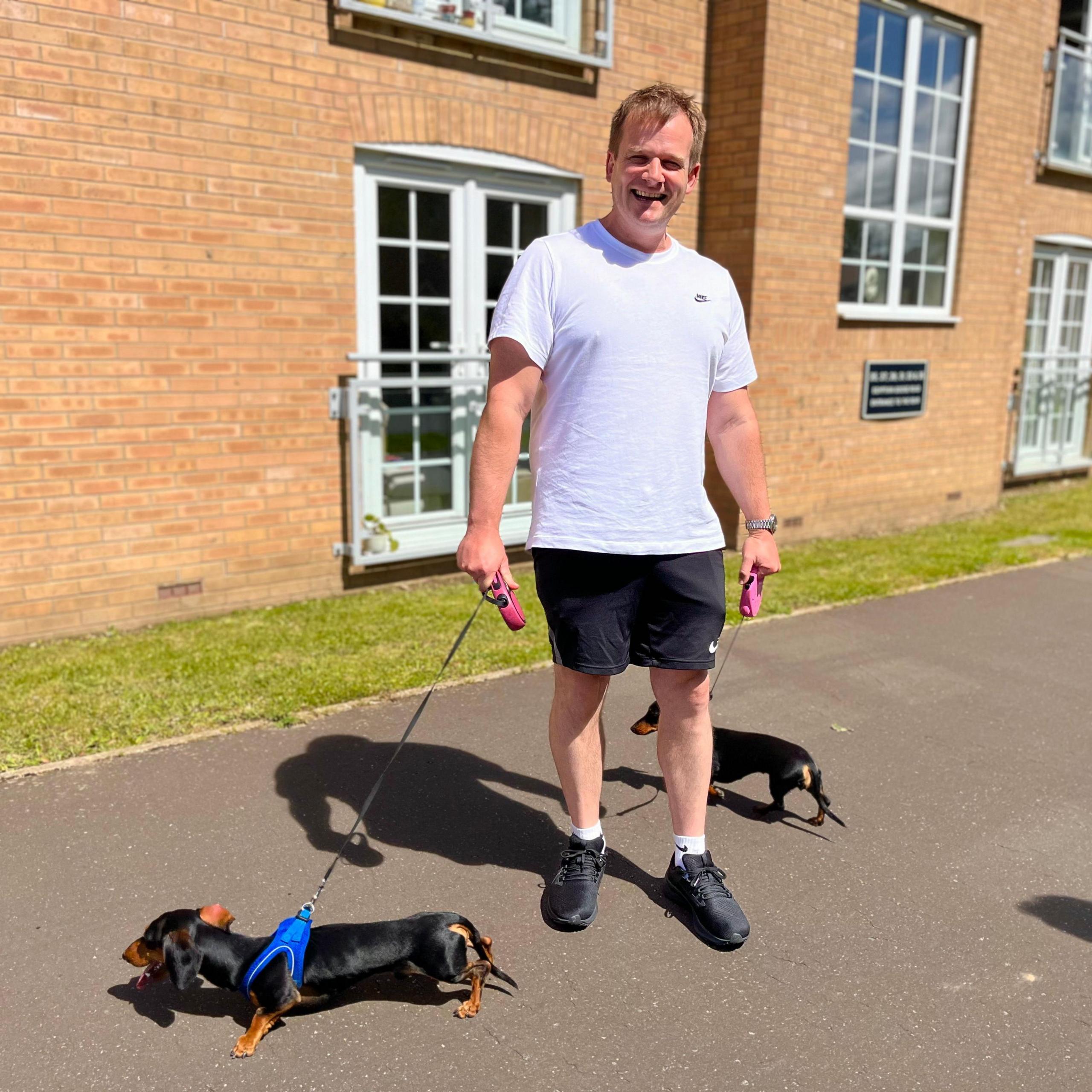 Stuart Johnson wears a white T-shirt and black shorts as he walks his two dachshunds