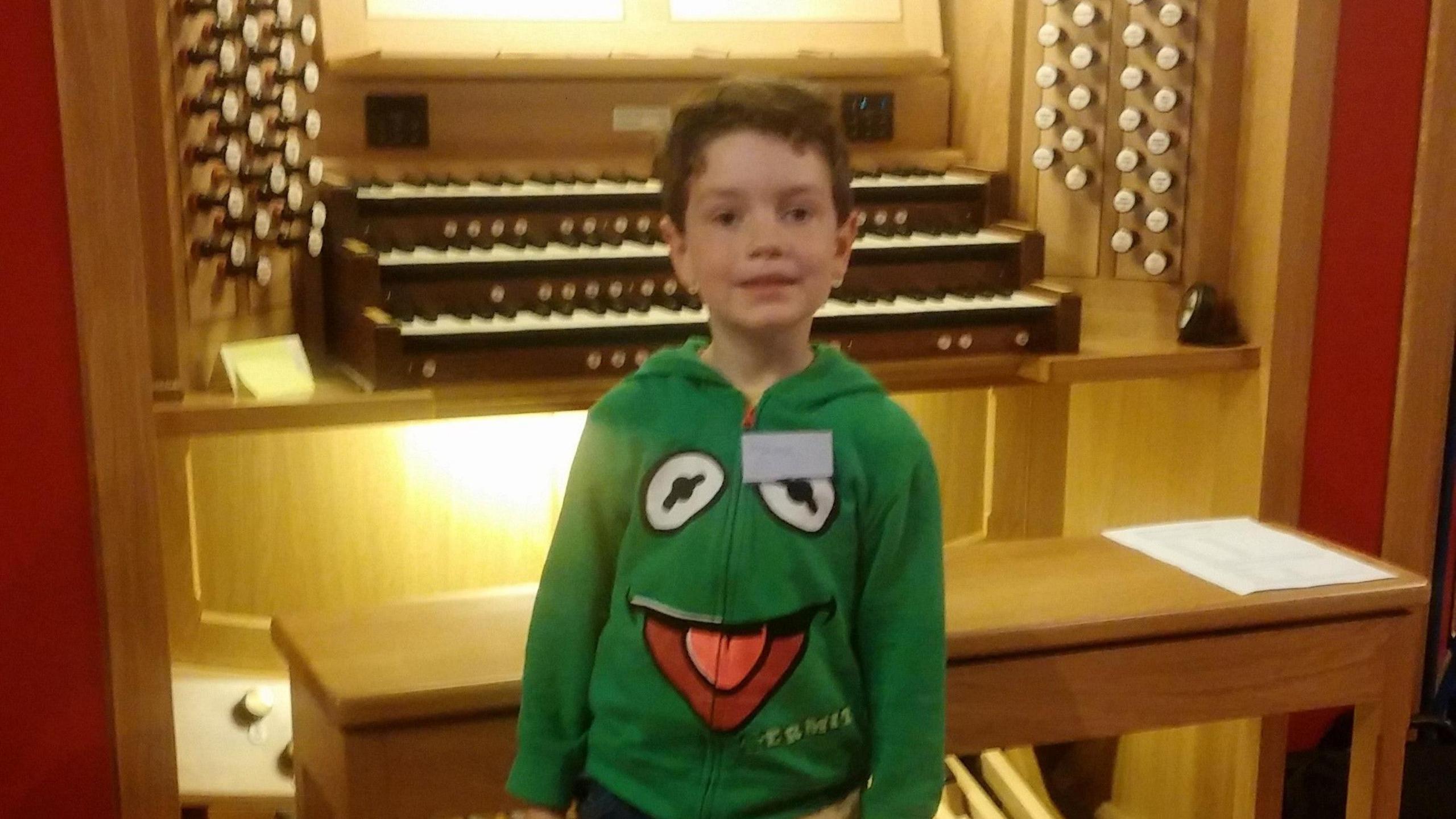 Six-year-old Llewelyn Blezard-Greenwell wearing a Kermit the Frog hoodie, standing in front of an organ.