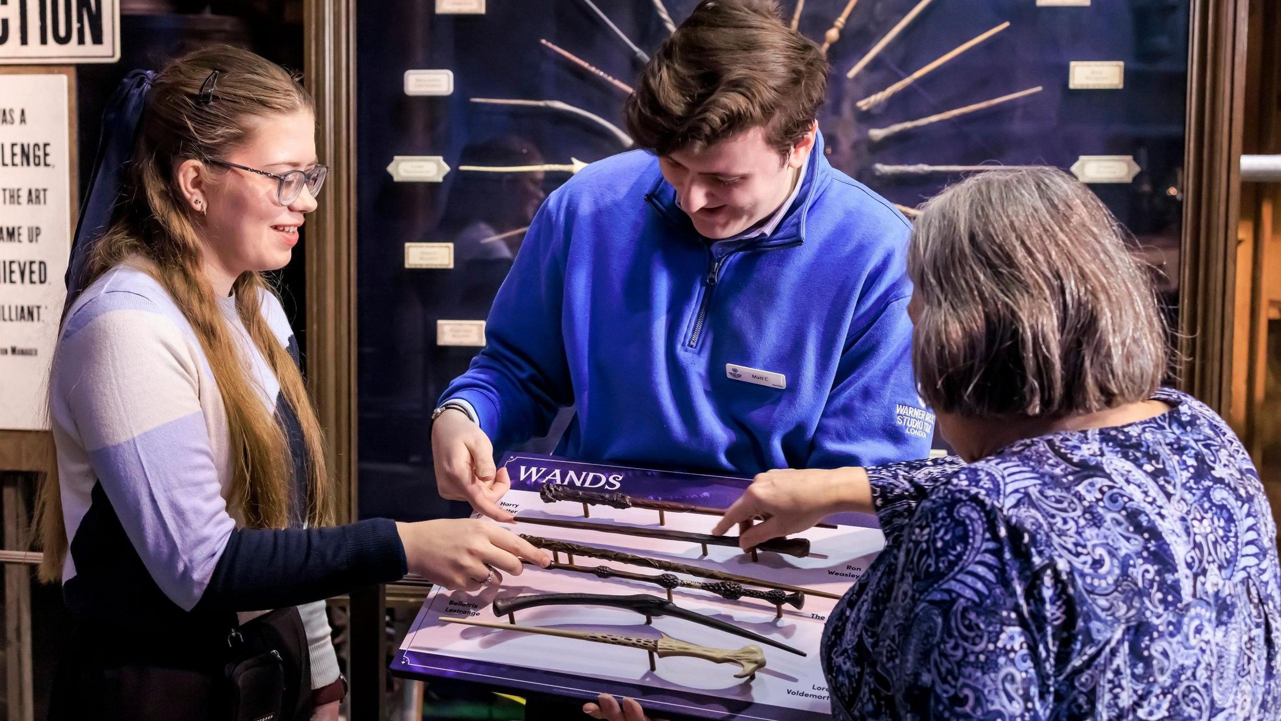 A young woman is reaching out to touch a wand prop that was used by Dumbledore in the Harry Potter films. It is among a selection of wands being held by a member of staff wearing a blue fleece. An older woman is reaching out to touch another wand in the selection. Behind them is a display of wands arranged in a circular pattern.