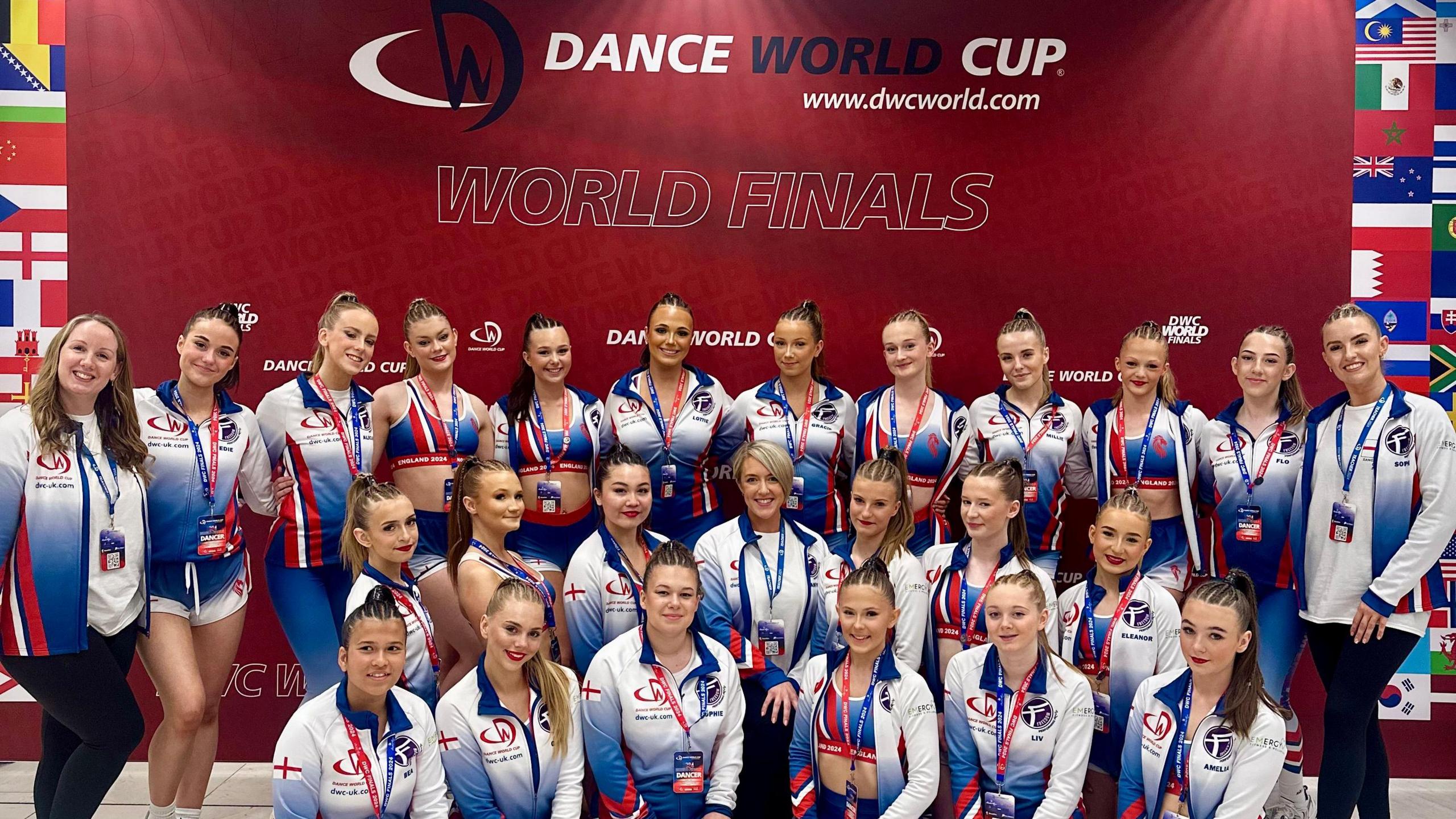 25 women shown standing in front of a sign saying 'Dance World Cup Finals'. All are wearing red white and blue jackets and have their hair braided. 

