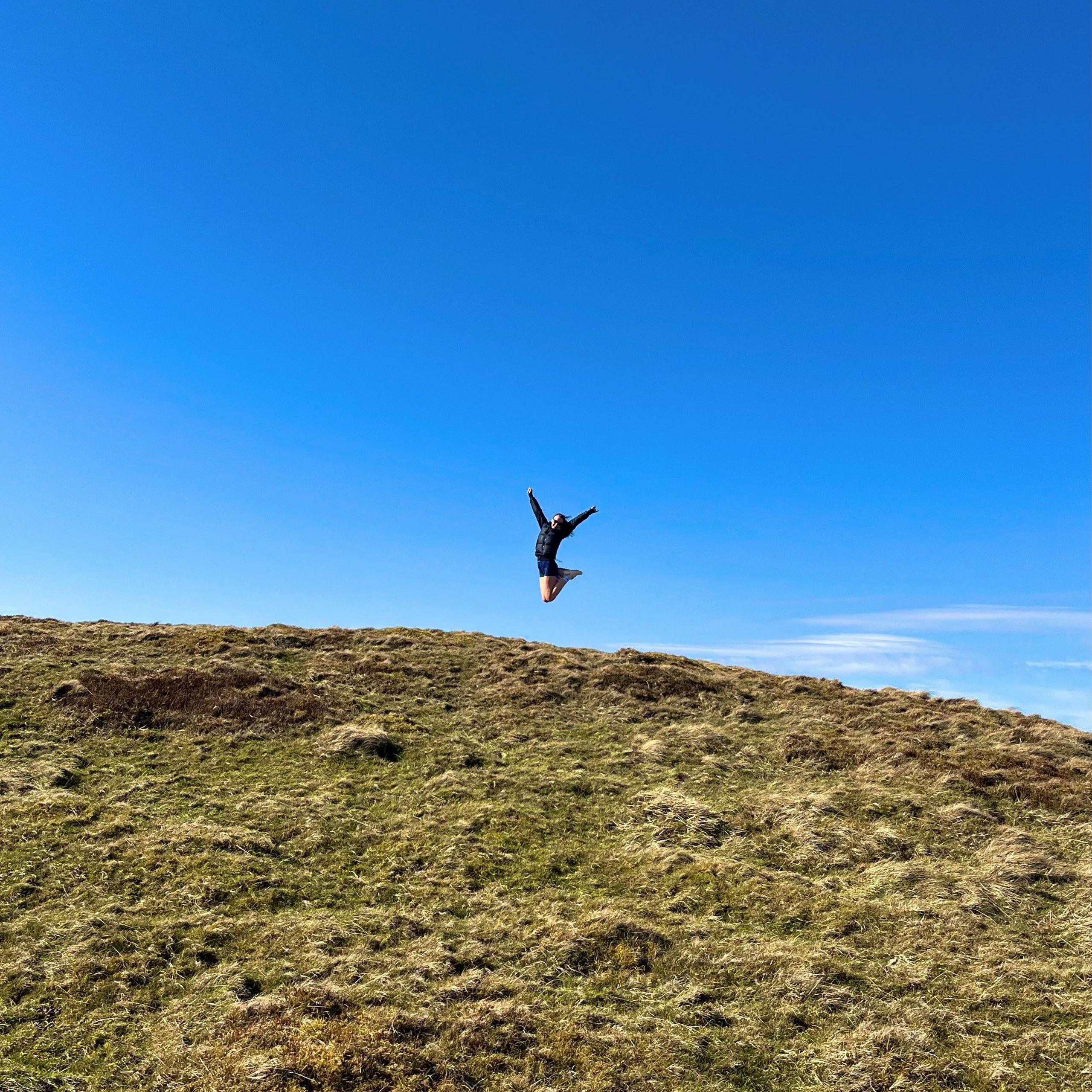 A woman jumps on the top of a hill. The grass in the foreground is green. The woman jumping in the middle is wearing dark clothing. The sky is blue.