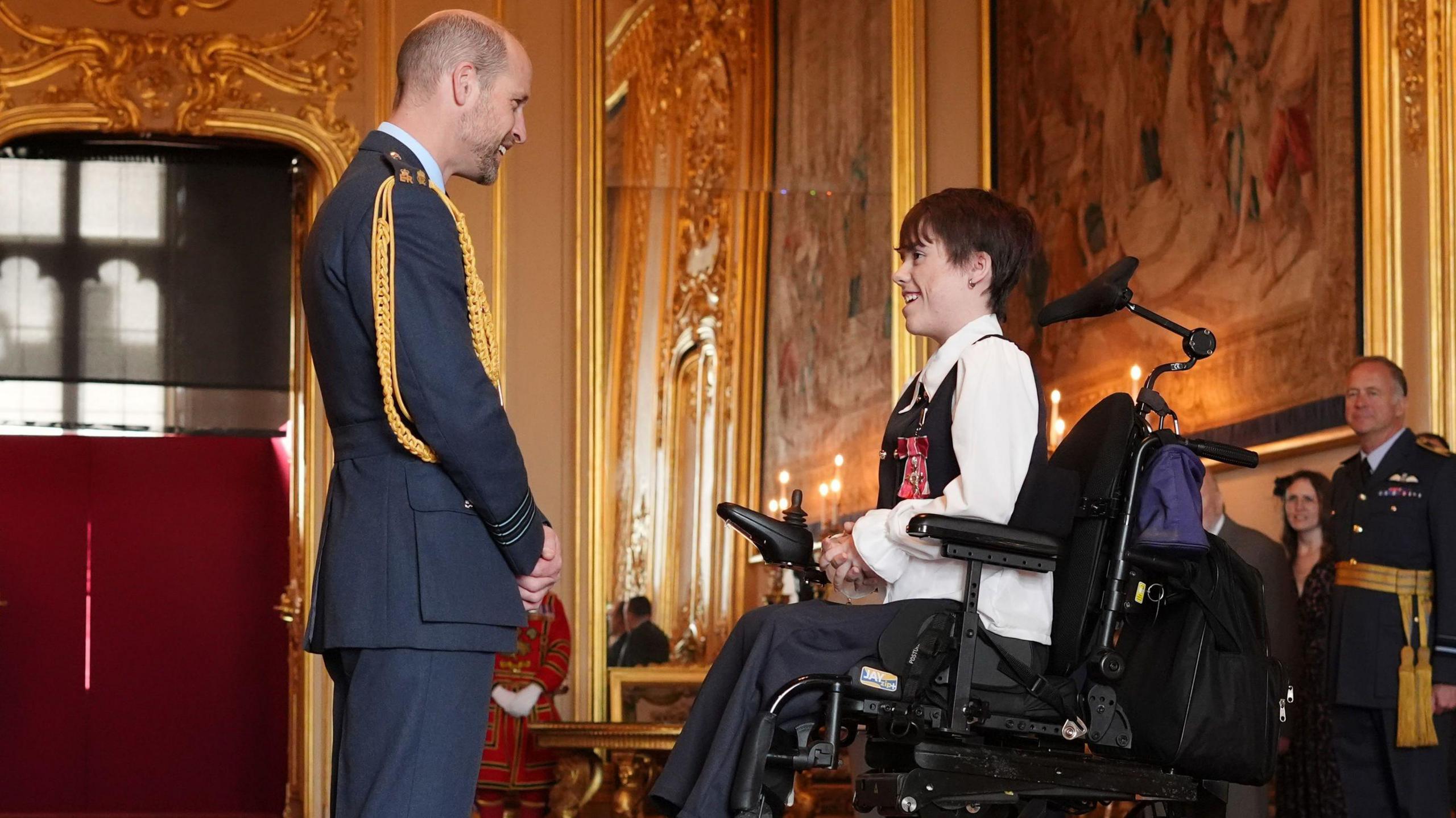 Carmela Chillery-Watson wearing a shirt and waist coat and her medal on her chest. She is sat in a specialist chair speaking to Prince William who is wearing military uniform at Windsor Castle.