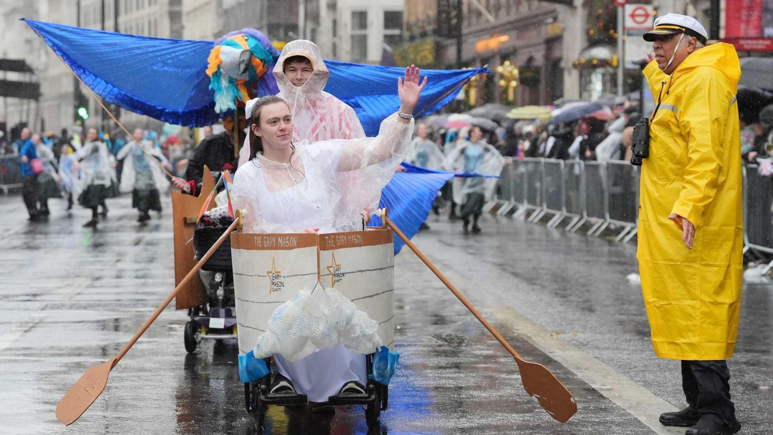 A woman sitting in a small boat on wheels with oars sticking out of the side. To the right is a man in a long yellow waterproof coat and a captain's hat. 