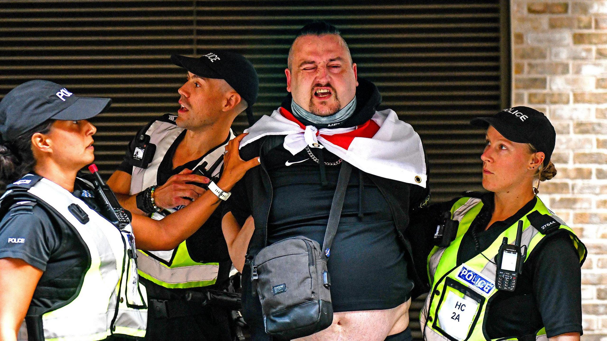 A shouting man wearing an England flag being held by three police officers