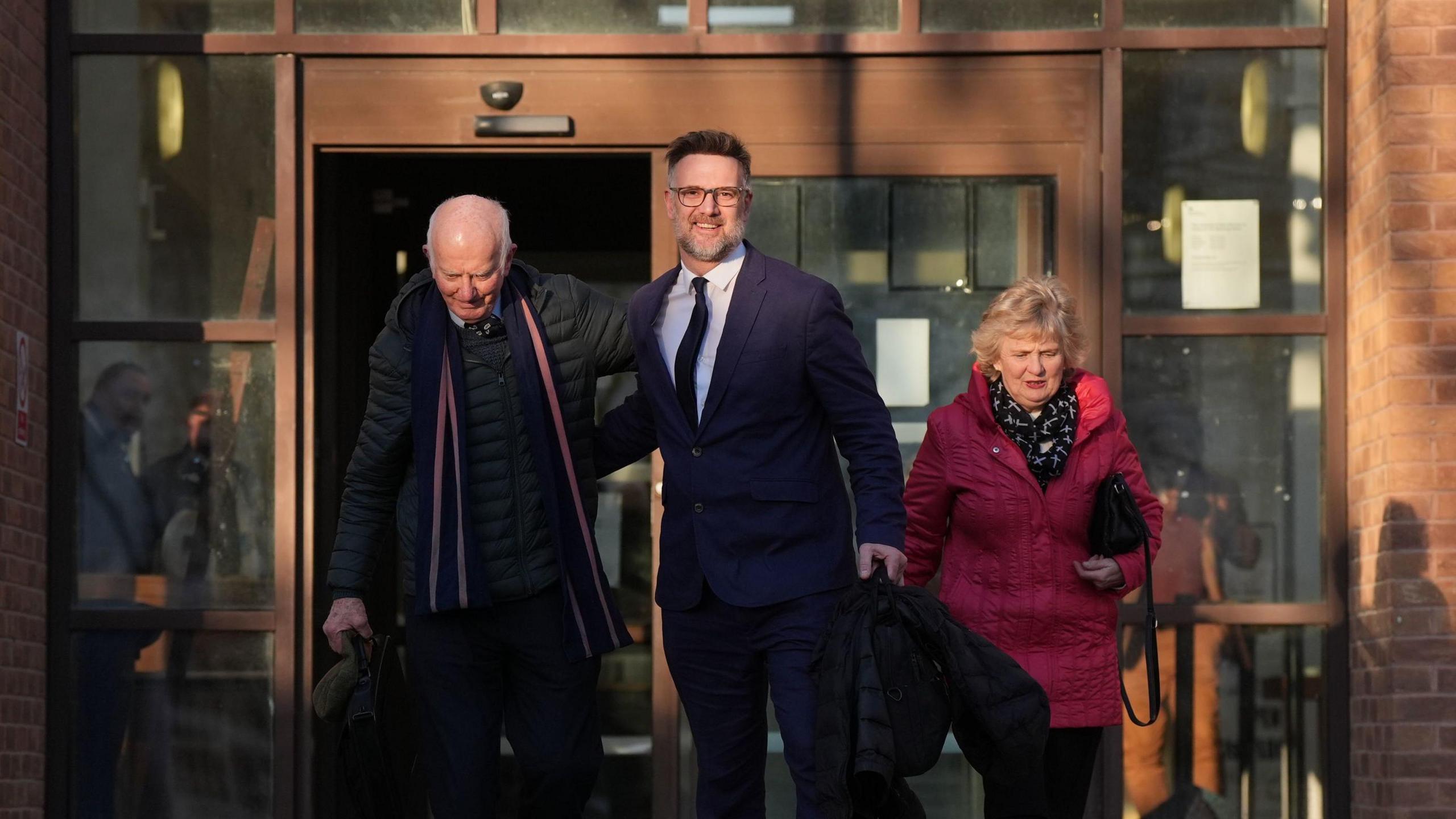 Charles Hanson pictured leaving the court with his parents. He is wearing a suit and tie. His dad is in a black puffer jacket with a scarf around his neck. His mum is in a red coat.