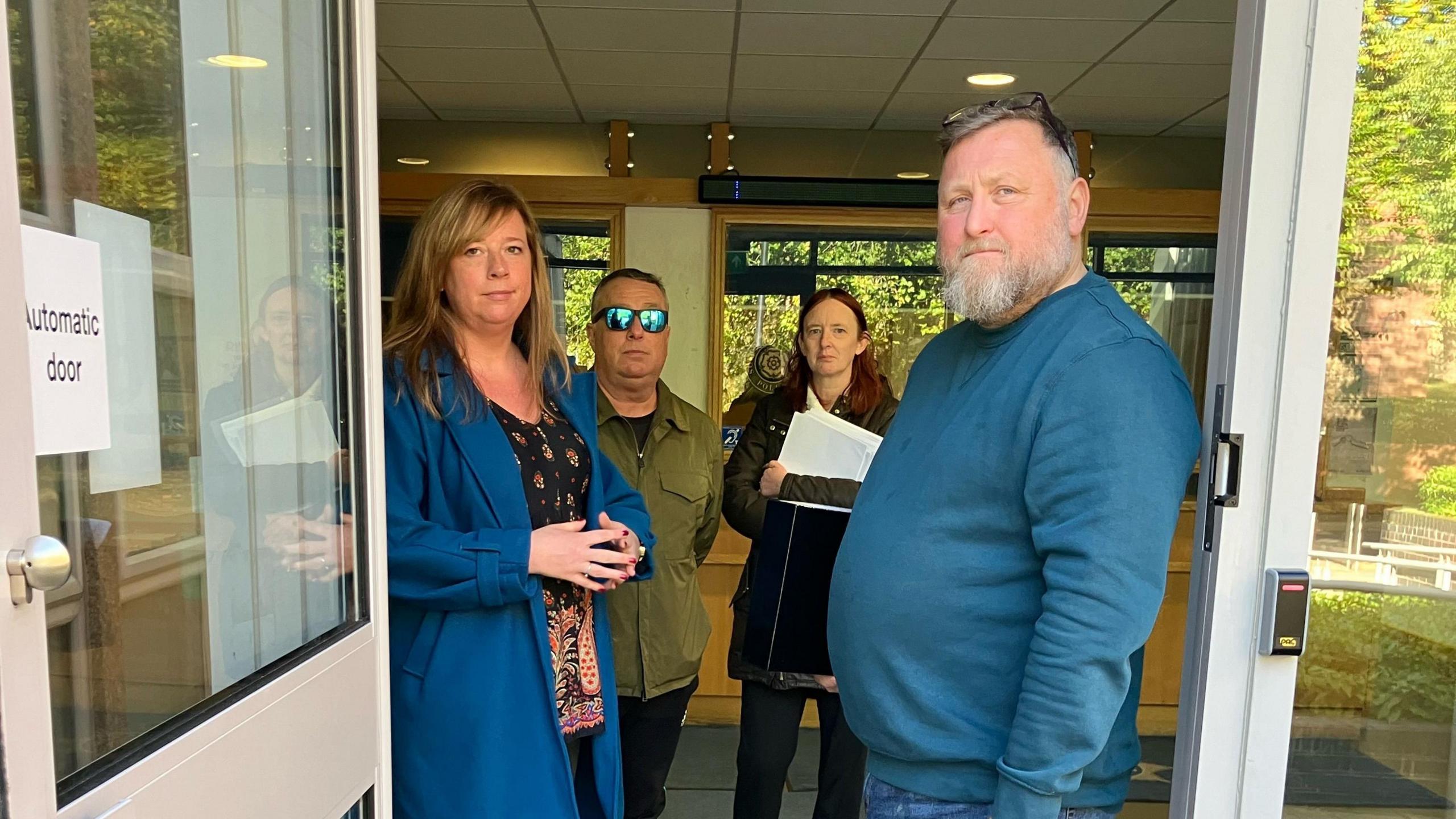 Four people standing in a doorway to a police station. One person is holding hundreds of pieces of paper