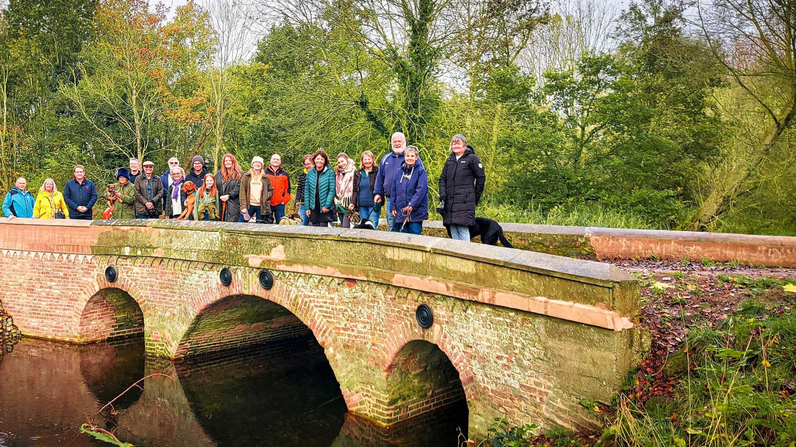 About 20 residents and council representatives standing in a line on Hempyard Bridge, looking at the camera. They are wearing coats, scarves and hats. One of them has a black dog. The brick-built bridge, which has three arches, is narrow and looks old. The bridge is over a river. There are trees behind it.