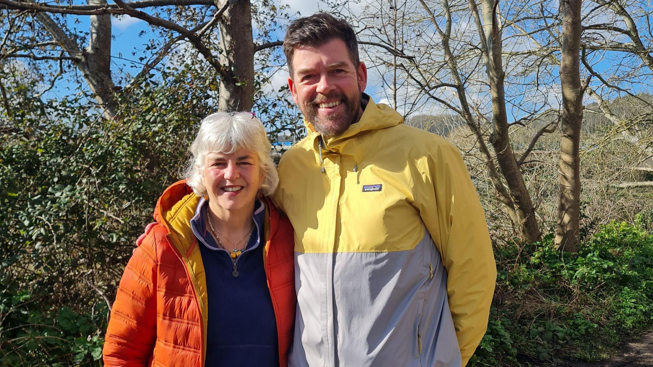 A man and a woman stand alongside each other looking at the camera. She has white hair and is wearing an orange padded jacket. He has a beard and is wearing a waterproof top which is a mixture of yellow and grey in colour. They are part of the Bristol Walk Fest