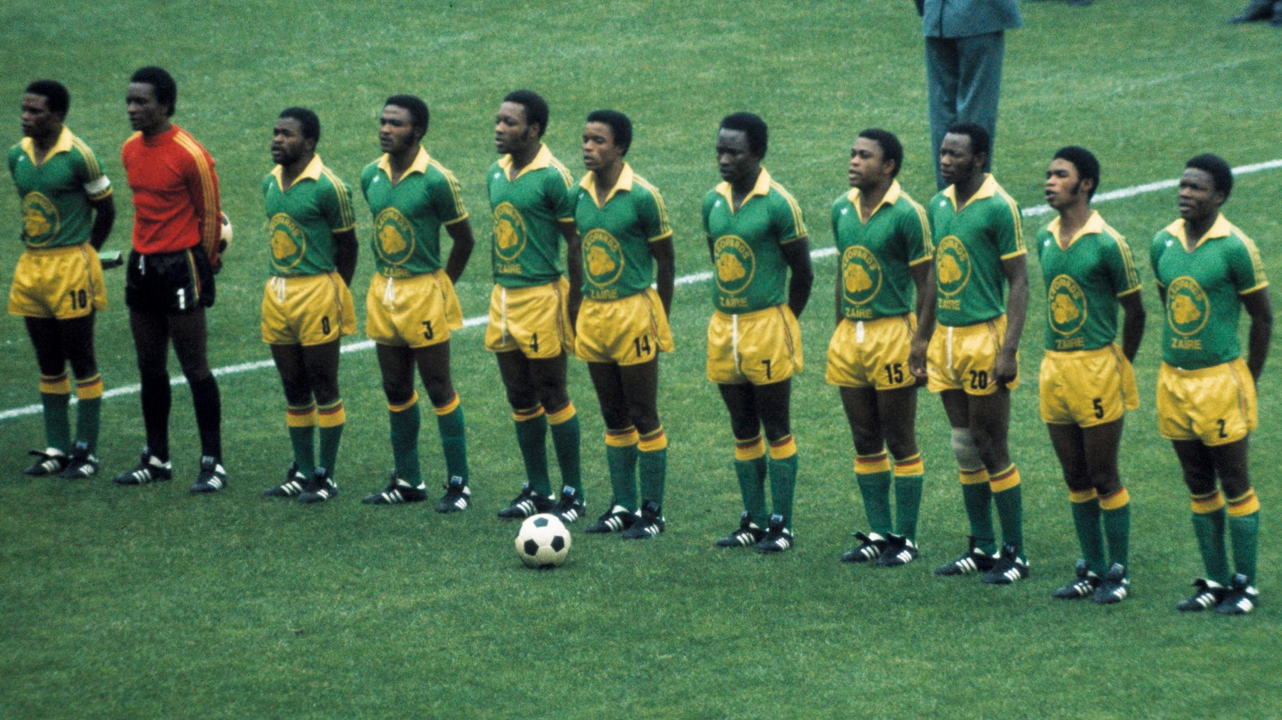 Zaire's football team line up for a team photo ahead of a match at the 1974 World Cup. The outfield players wear green shirts with the image of a leopard on the chest, yellow shorts, green socks and black boots. The goalkeeper wears a red long-sleeved jersey, black shorts and black socks