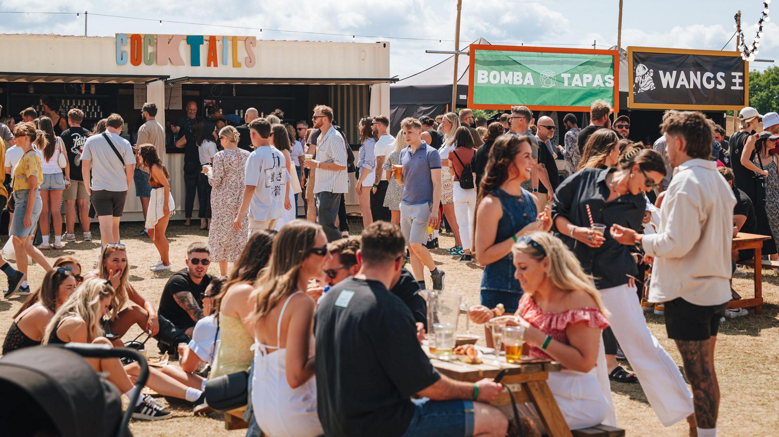 Large groups of people sit at the centre of the Feast On Bristol event on The Downs. It is a sunny day and they are wearing light summer clothing. The ground is dry and beige in colour after several weeks of dry weather. Around the edge of the open space various food stalls are visible.