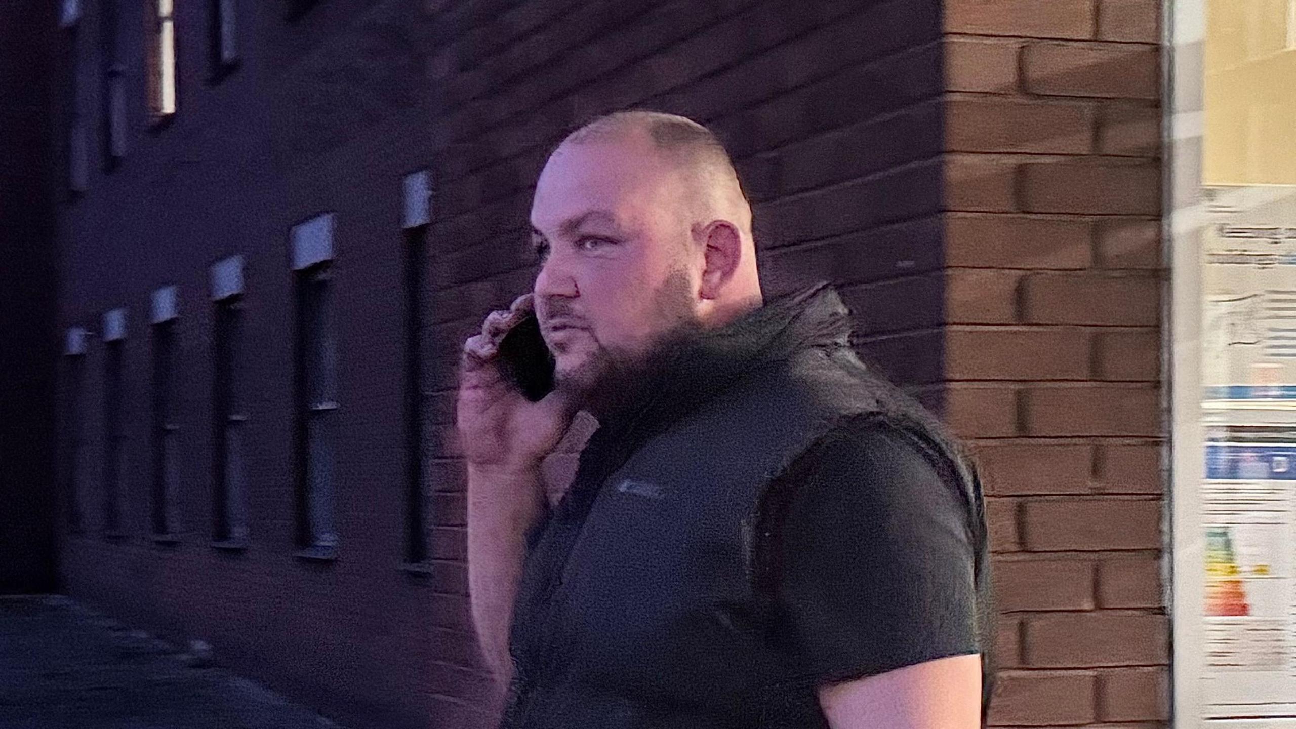 Adam Fox - a man wearing a black Gillet over a black t shirt walking out of court while on the phone.