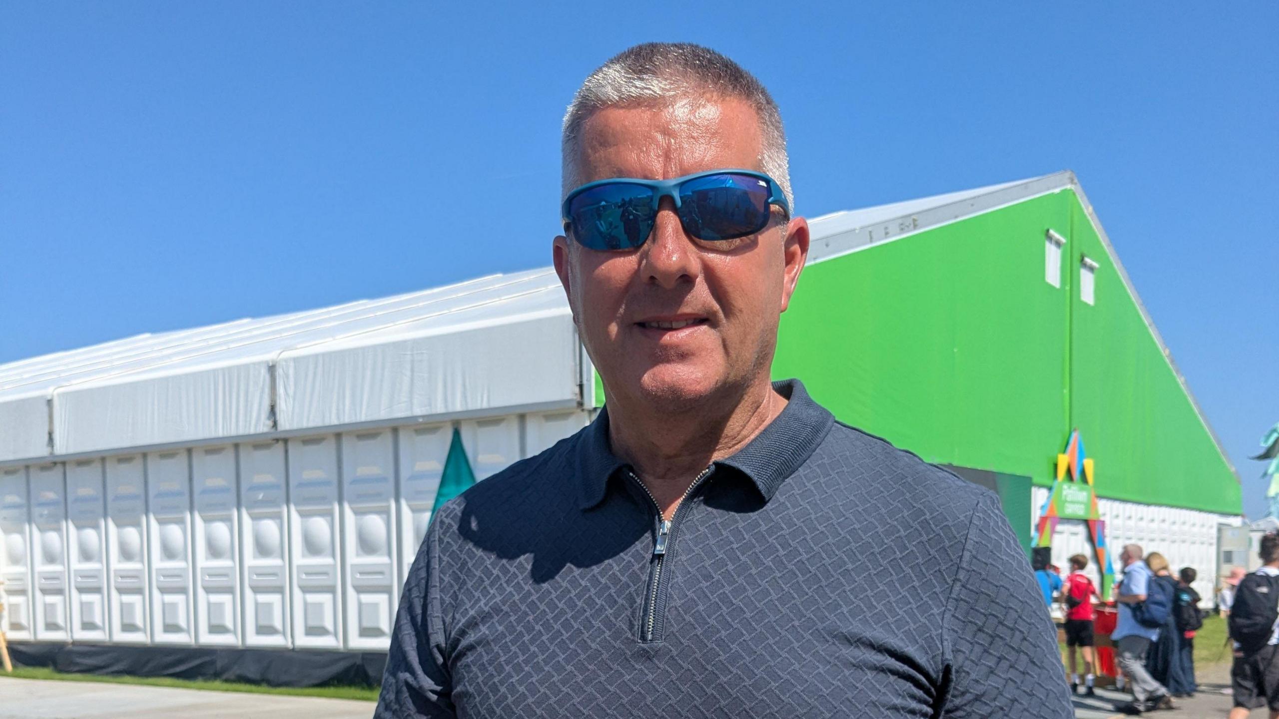 Llun o Clive Williams, pennaeth Ysgol Gyfun Penweddig yn sefyll ar y maes gyda sbectol haul. 